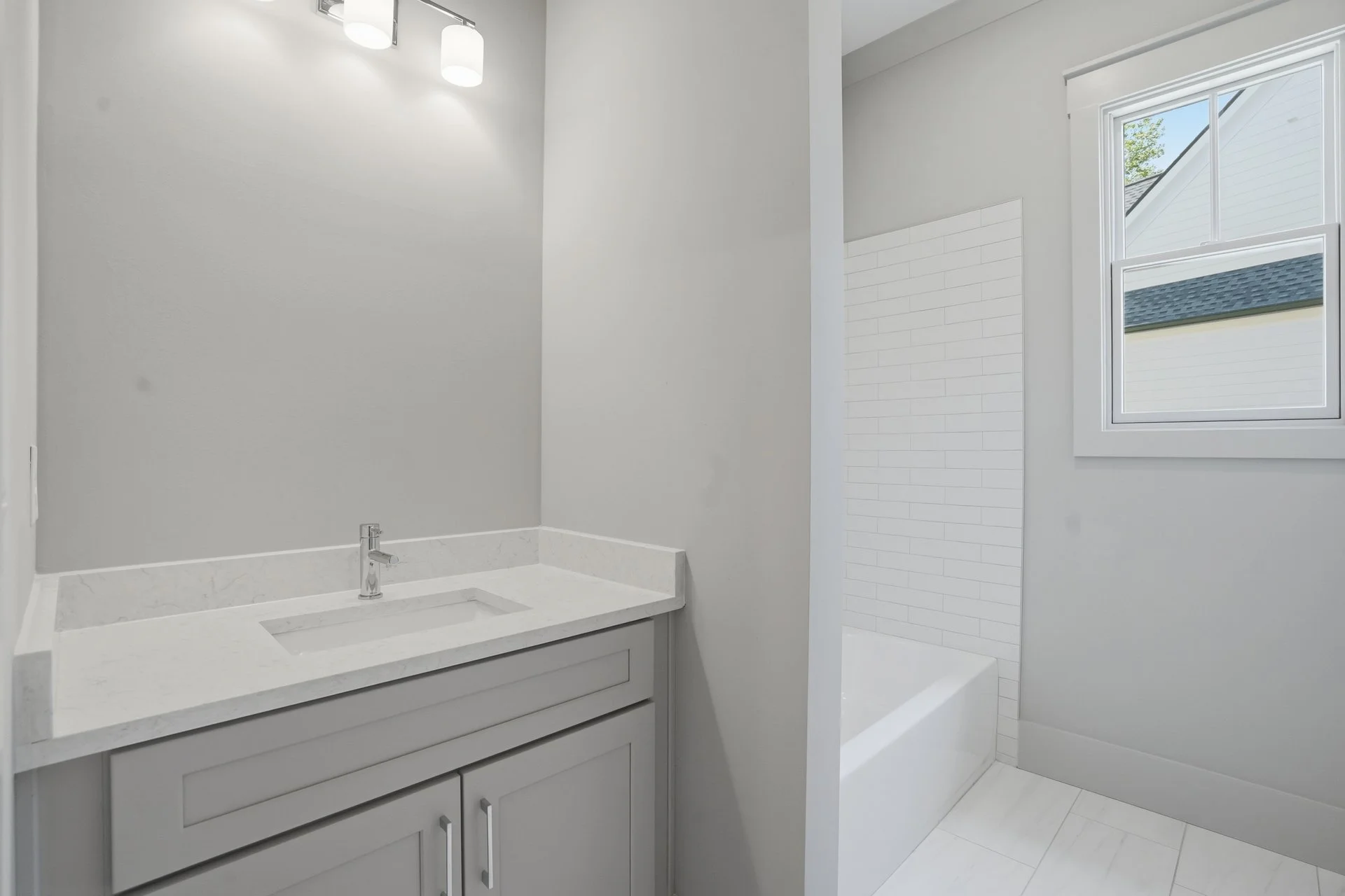 Modern bathroom with white sink countertop, gray cabinetry, a wall-mounted faucet, white tile flooring, and a shower area with a window.