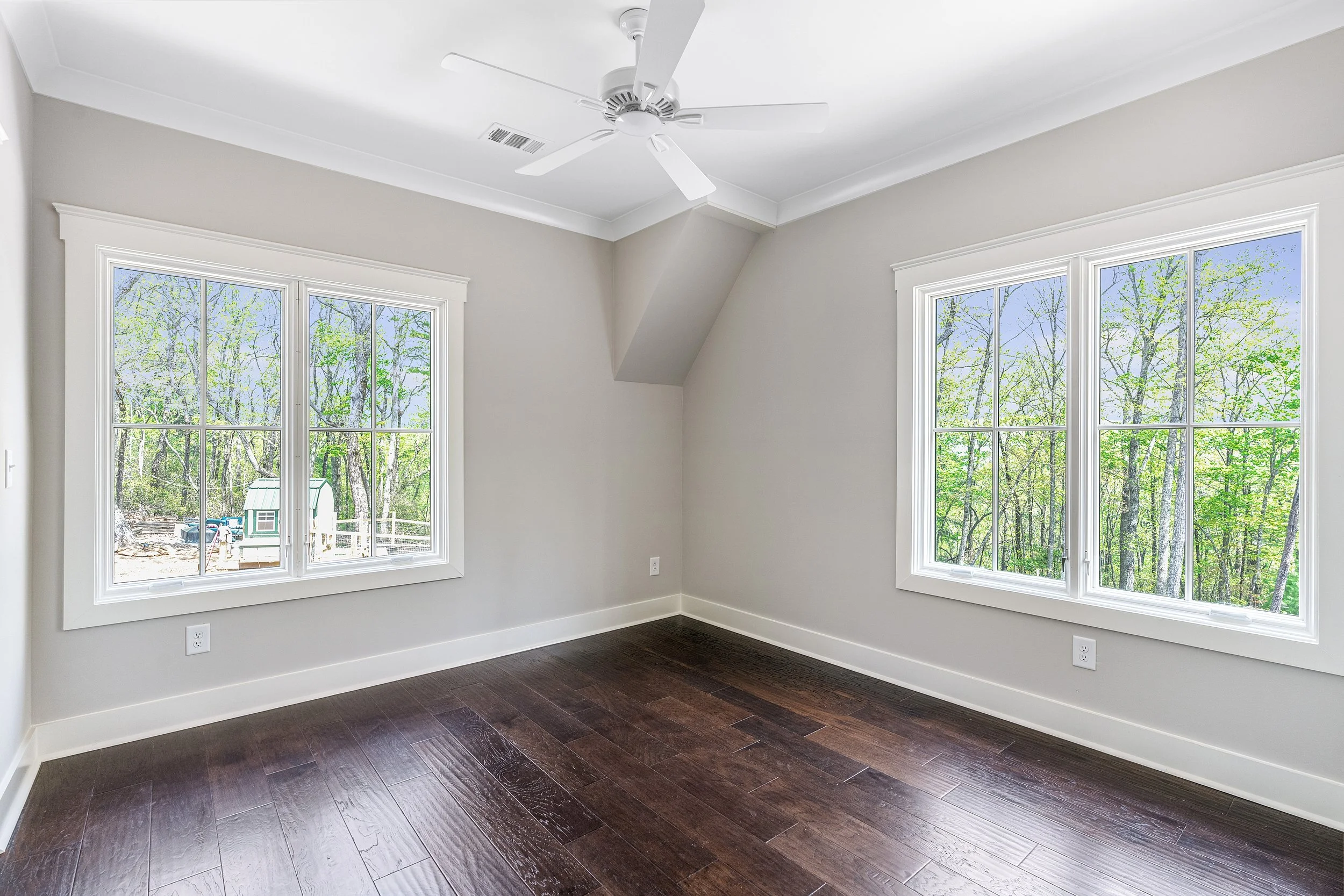 Empty room with large windows showing a wooded outdoor view, a ceiling fan, and dark hardwood flooring.