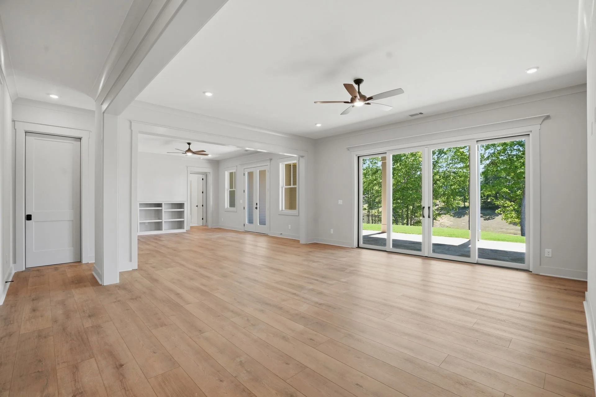 Empty living room with light hardwood floors, white walls, ceiling fans, large glass sliding doors showing a backyard with green trees, and windows with white trim.