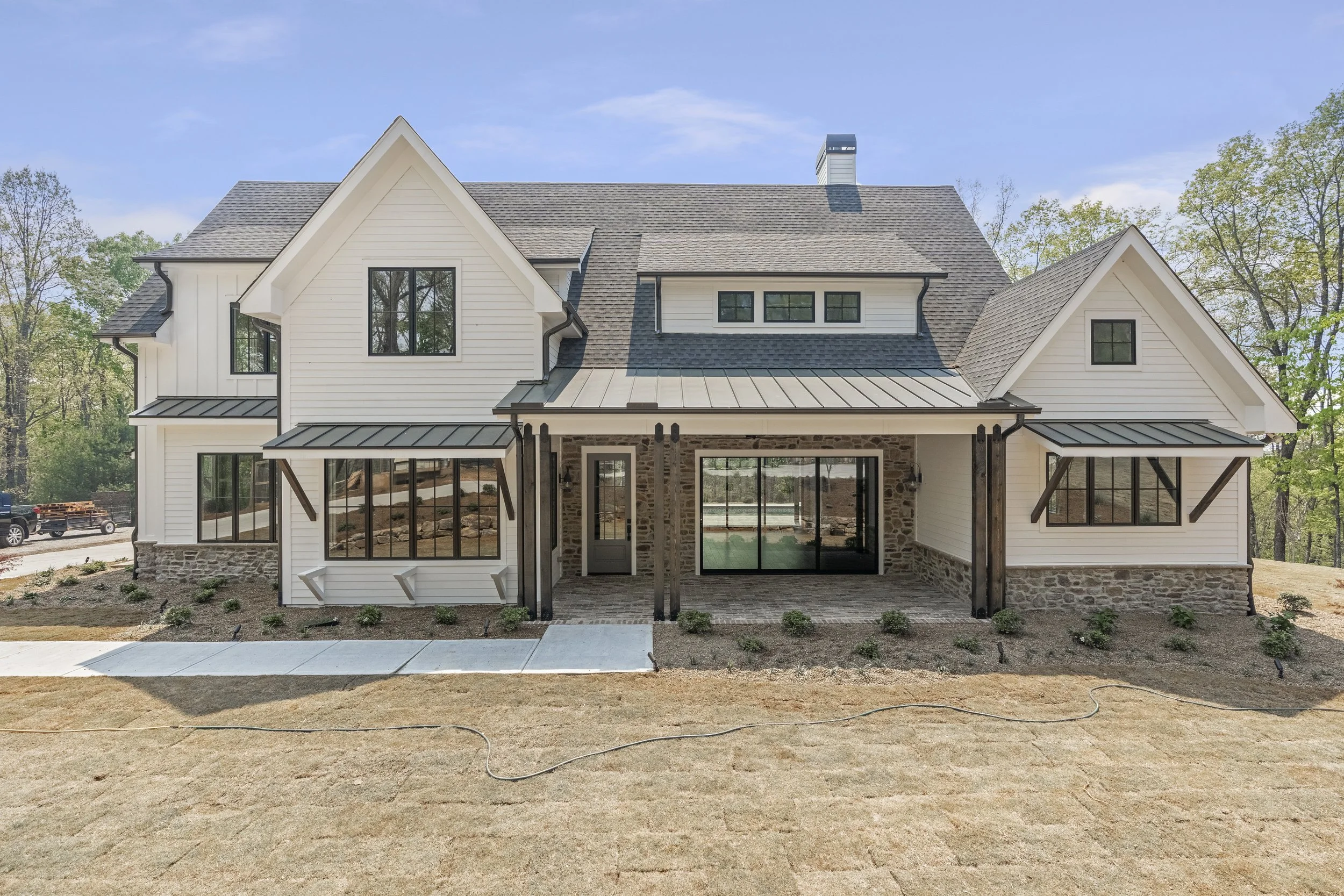 Newly constructed two-story house with white siding, a stone foundation, and black-framed windows. The front entrance is framed with a small porch, and the backyard features sliding glass doors. The house has a complex roof with multiple gables and d
