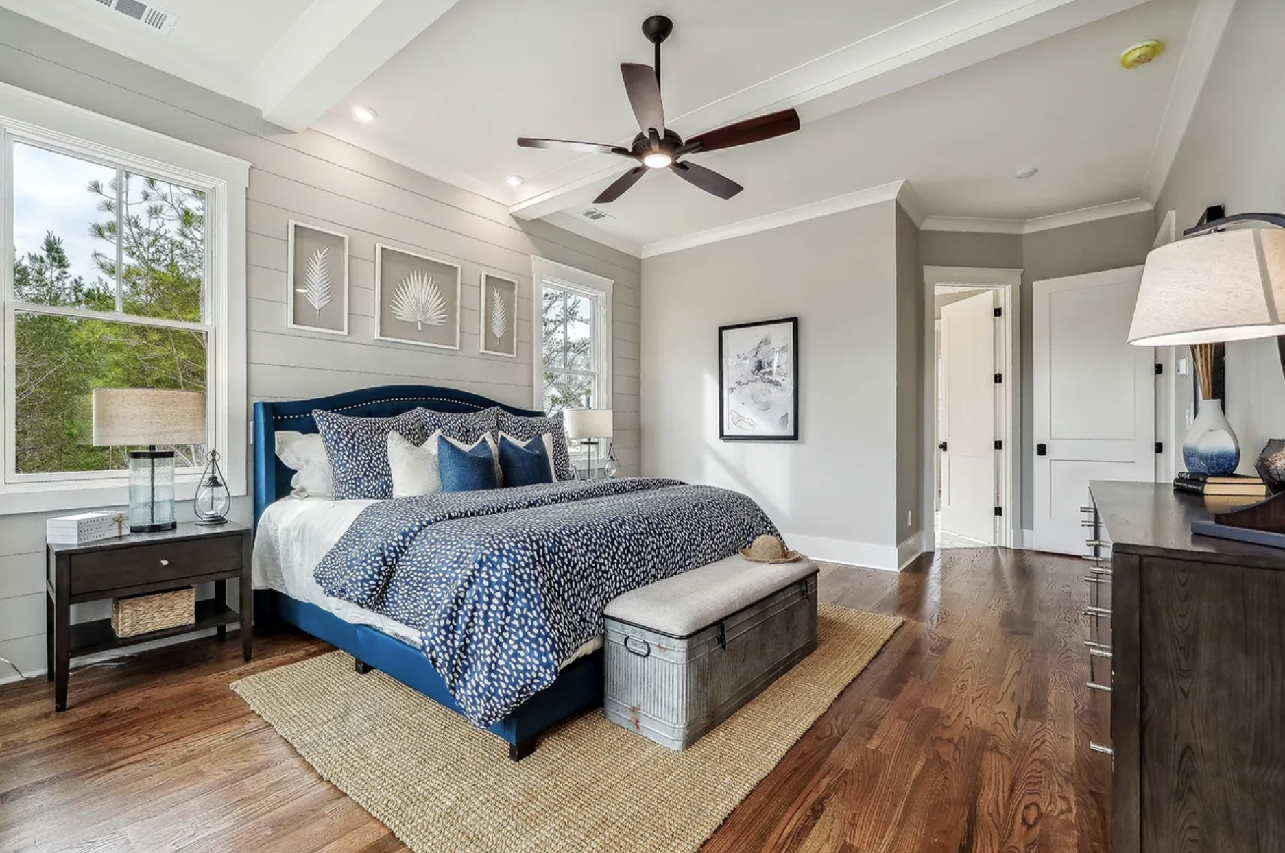 A bedroom with a bed featuring blue and white bedding, a dark wood nightstand with a lamp, framed artwork on the wall, windows with trees outside, a ceiling fan, a dresser with a table lamp, and a woven rug on hardwood floor.