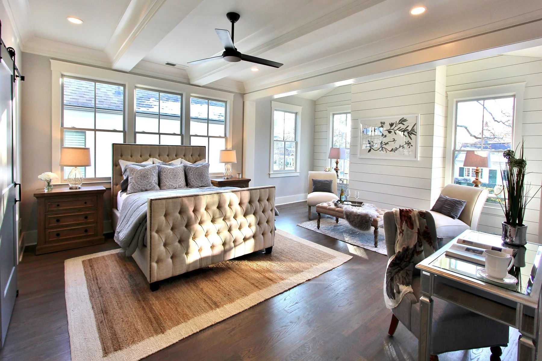 A bedroom with a beige tufted bed, wooden nightstands with lamps, and large windows letting in natural light. There are chairs, a small window, and a decorative wall art piece, with hardwood floors and a beige area rug.