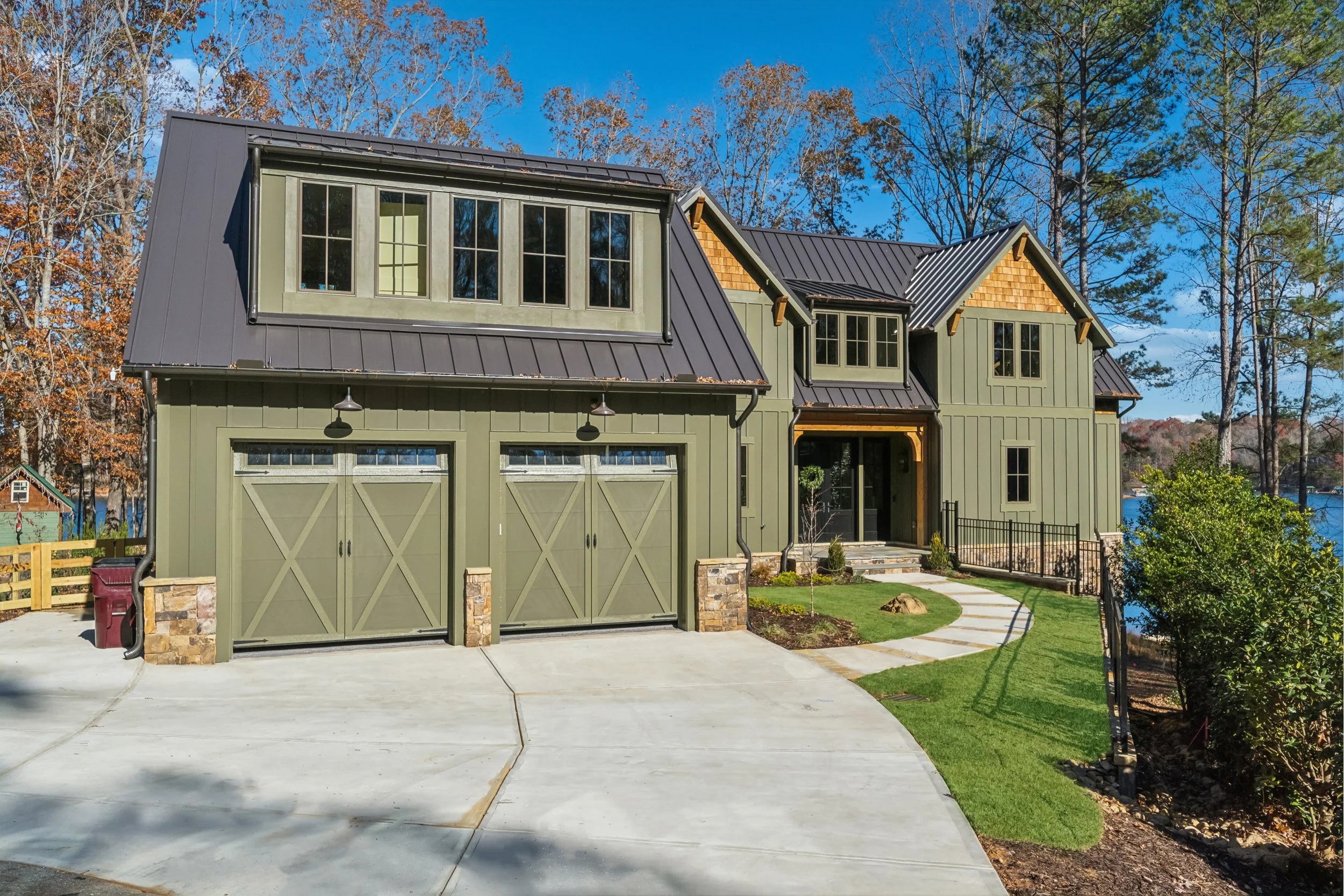 Front view of a modern house with a green exterior, a two-car garage, a manicured lawn, and tall trees in the background under a bright blue sky.