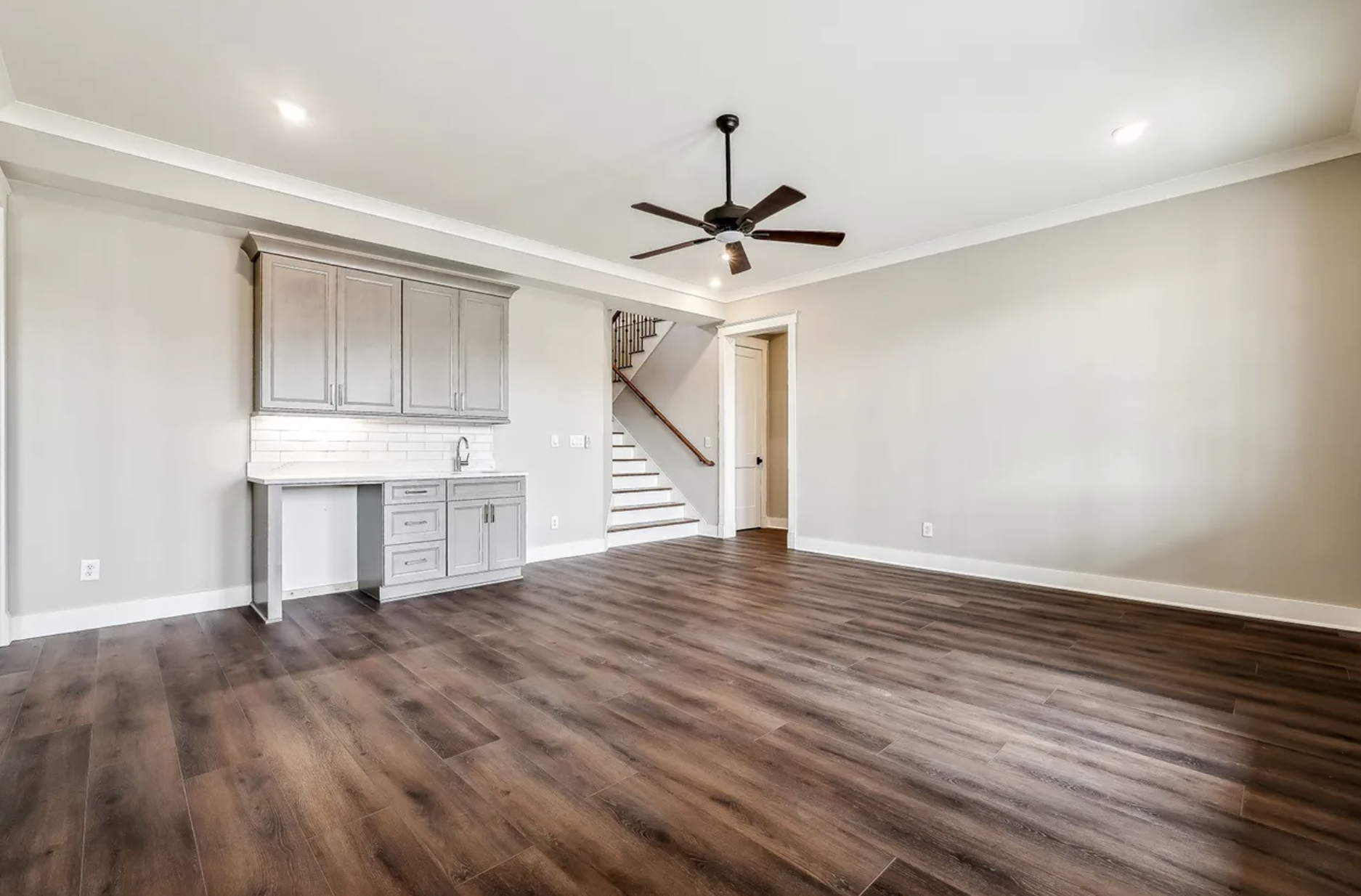 Empty living room with hardwood floors, white walls, a ceiling fan, a staircase, and a built-in cabinet with a small sink area.