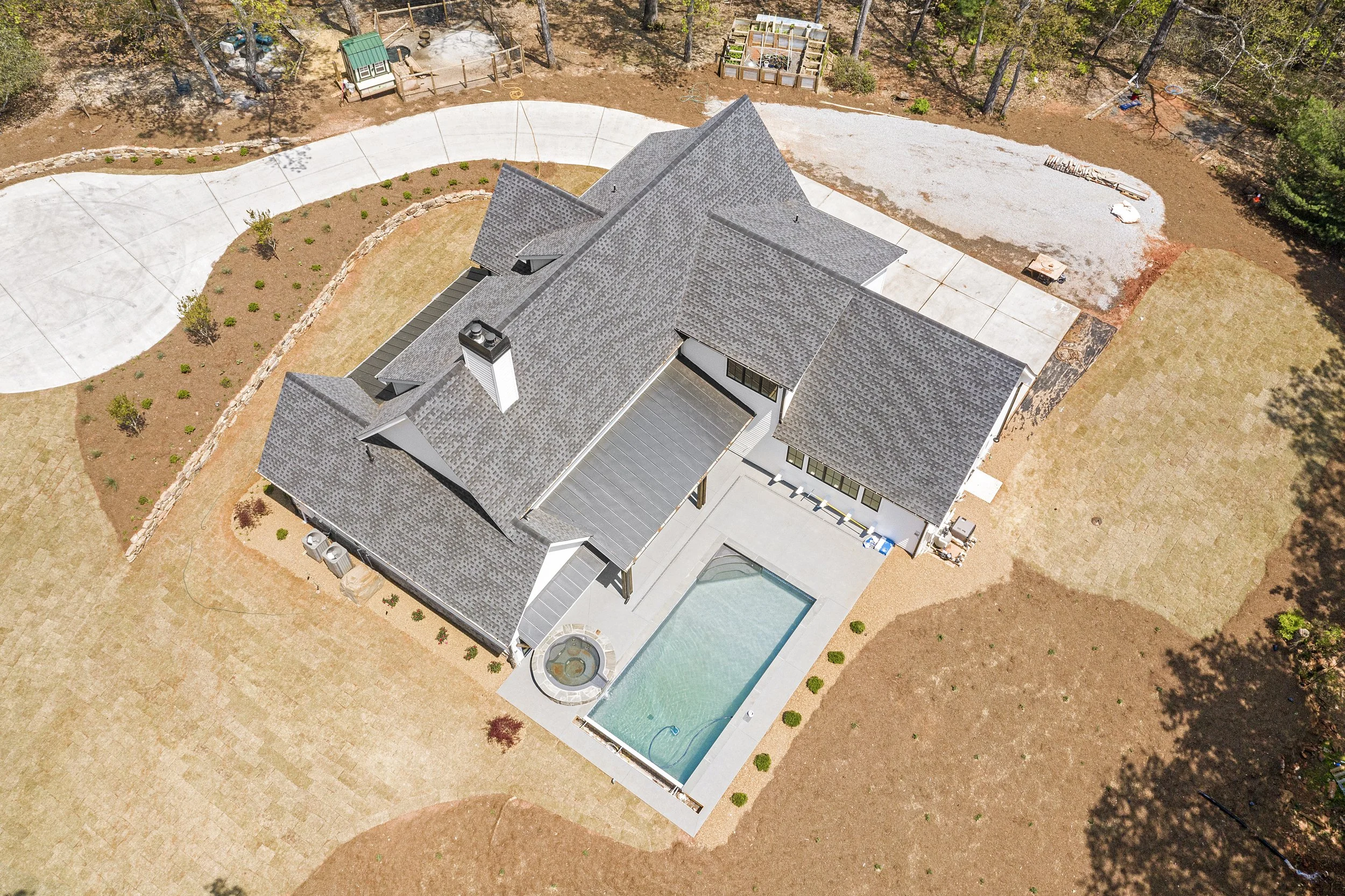 Aerial view of a modern house with a gray shingled roof, a swimming pool, and a surrounding yard with small trees, gravel areas, and a paved driveway.