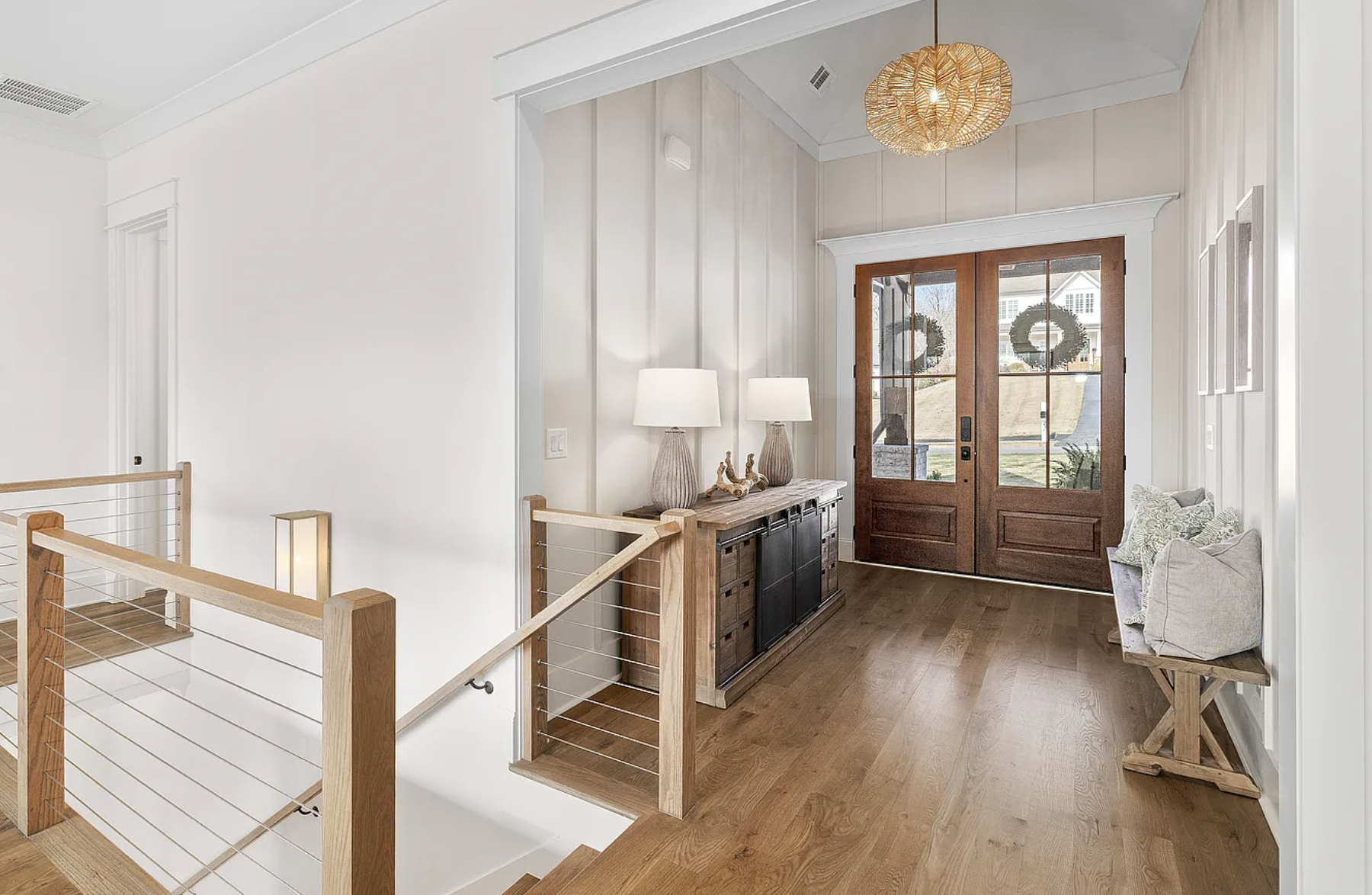 Entryway with wooden double doors decorated with wreaths, a console table with lamp and decorative driftwood, a cushioned bench with pillows, and a modern chandelier, featuring light wood flooring and white walls.