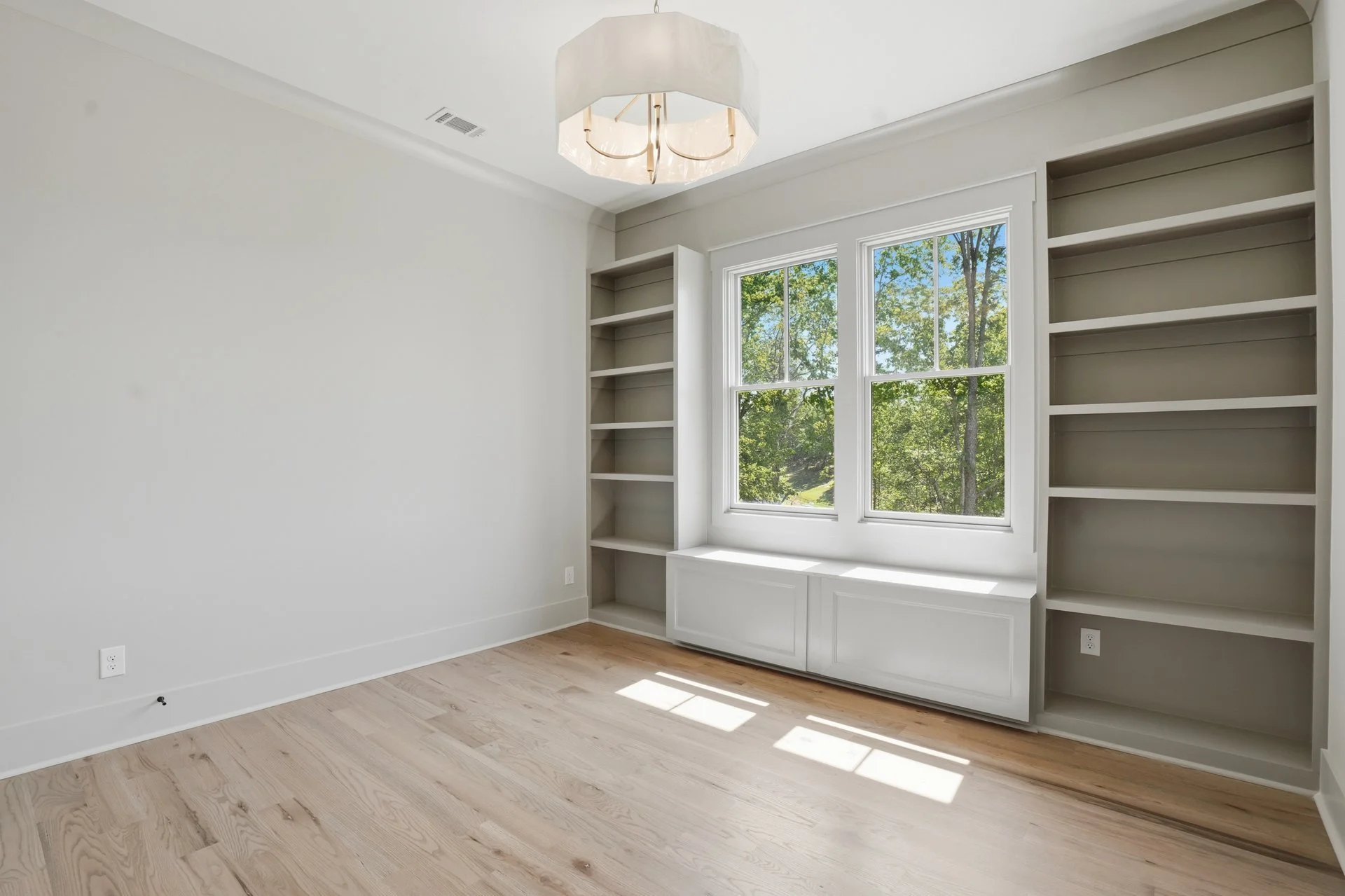 Empty white room with built-in shelves on both sides of the window, hardwood flooring, and a ceiling light fixture, with visible natural light coming through the window showing trees outside.
