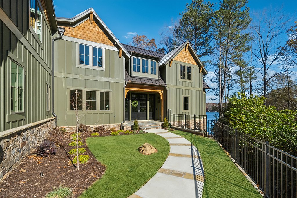 Front view of a two-story house with green siding and stone accents, surrounded by landscaped yard and tall trees, with a lake visible in the background.