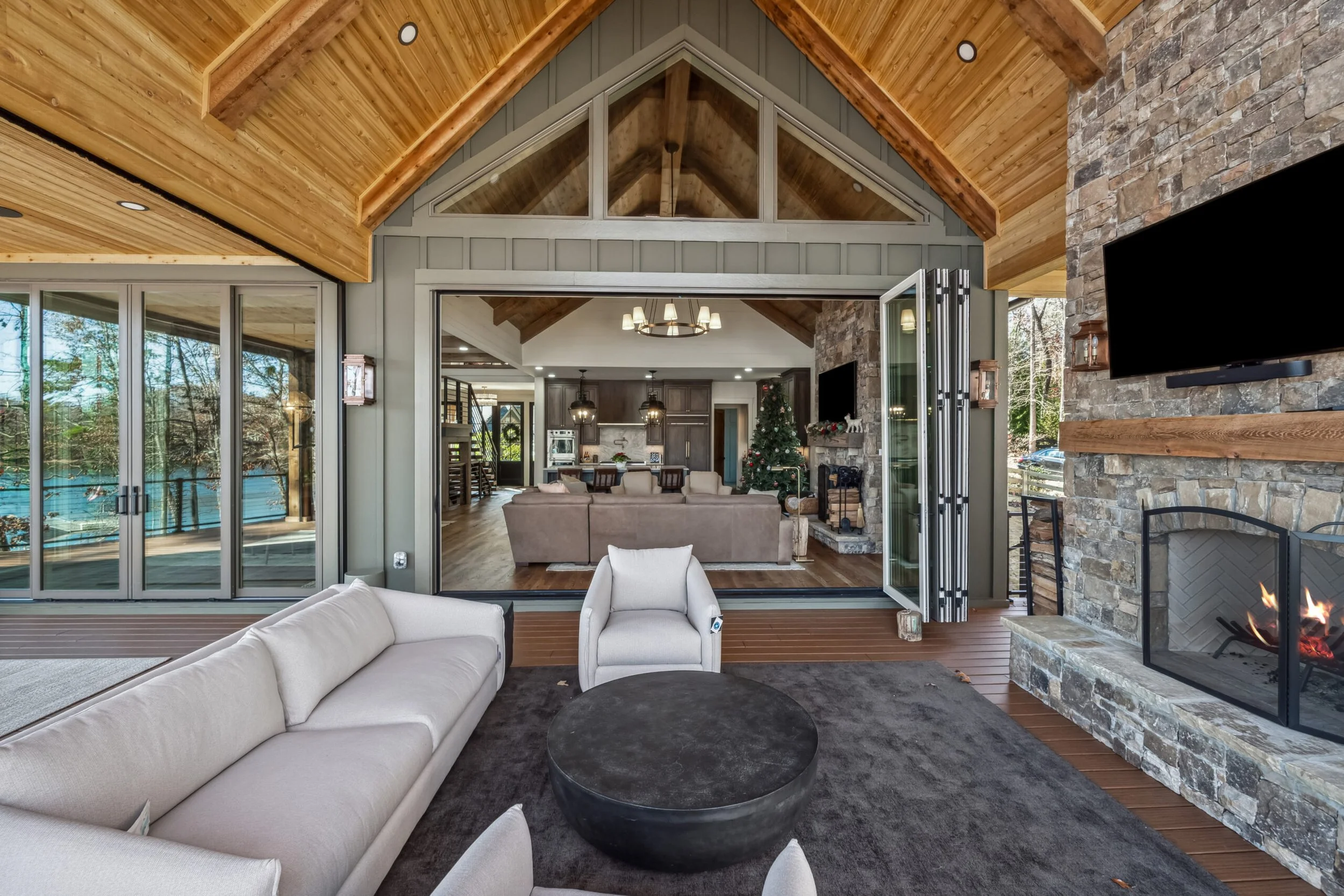 Open-concept living room with a stone fireplace, beige sofa, and large sliding glass doors opening to an outdoor deck, with a Christmas tree visible in the background.