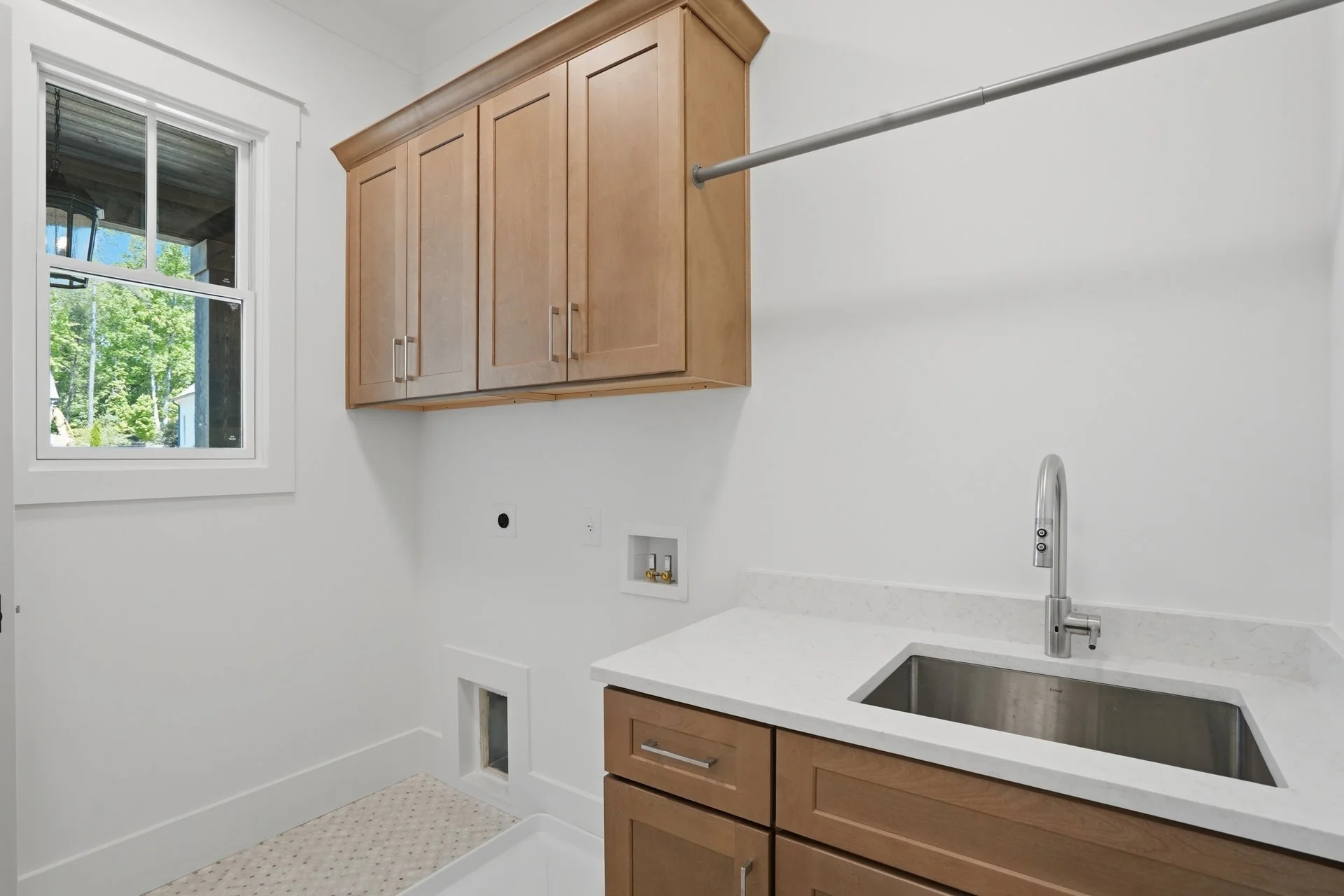 A laundry room with a window, wooden upper cabinet, white walls, a sink with a faucet, and a wall outlet.