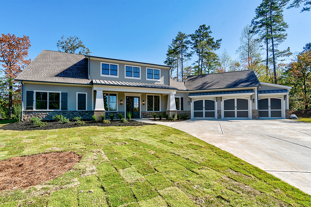 Modern two-story house with a gray roof, three-car garage, and front lawn under a clear blue sky, surrounded by trees.
