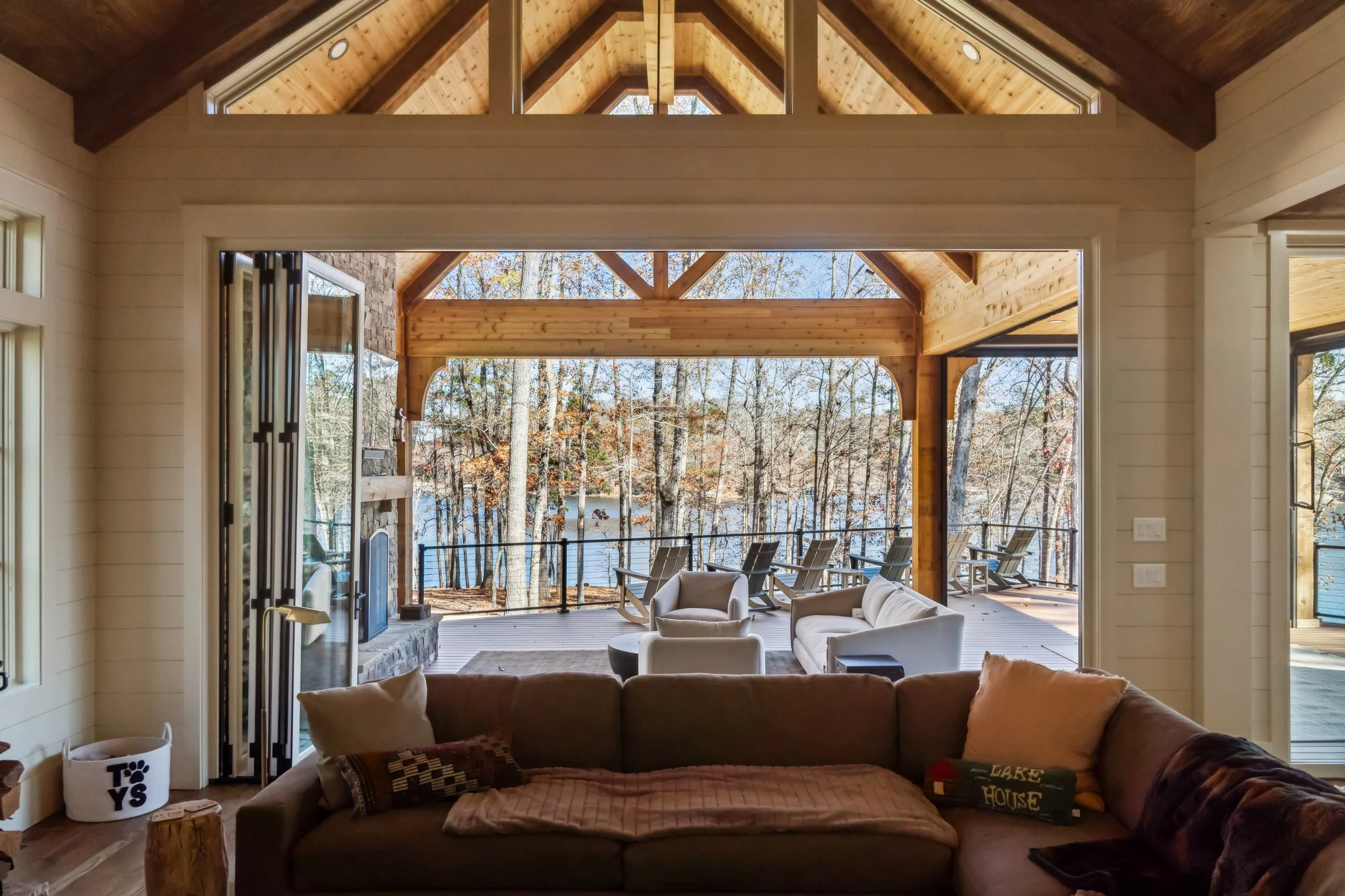 Living room with large open sliding doors leading to an outdoor deck overlooking a wooded lake area, with indoor seating including a sofa and chairs.