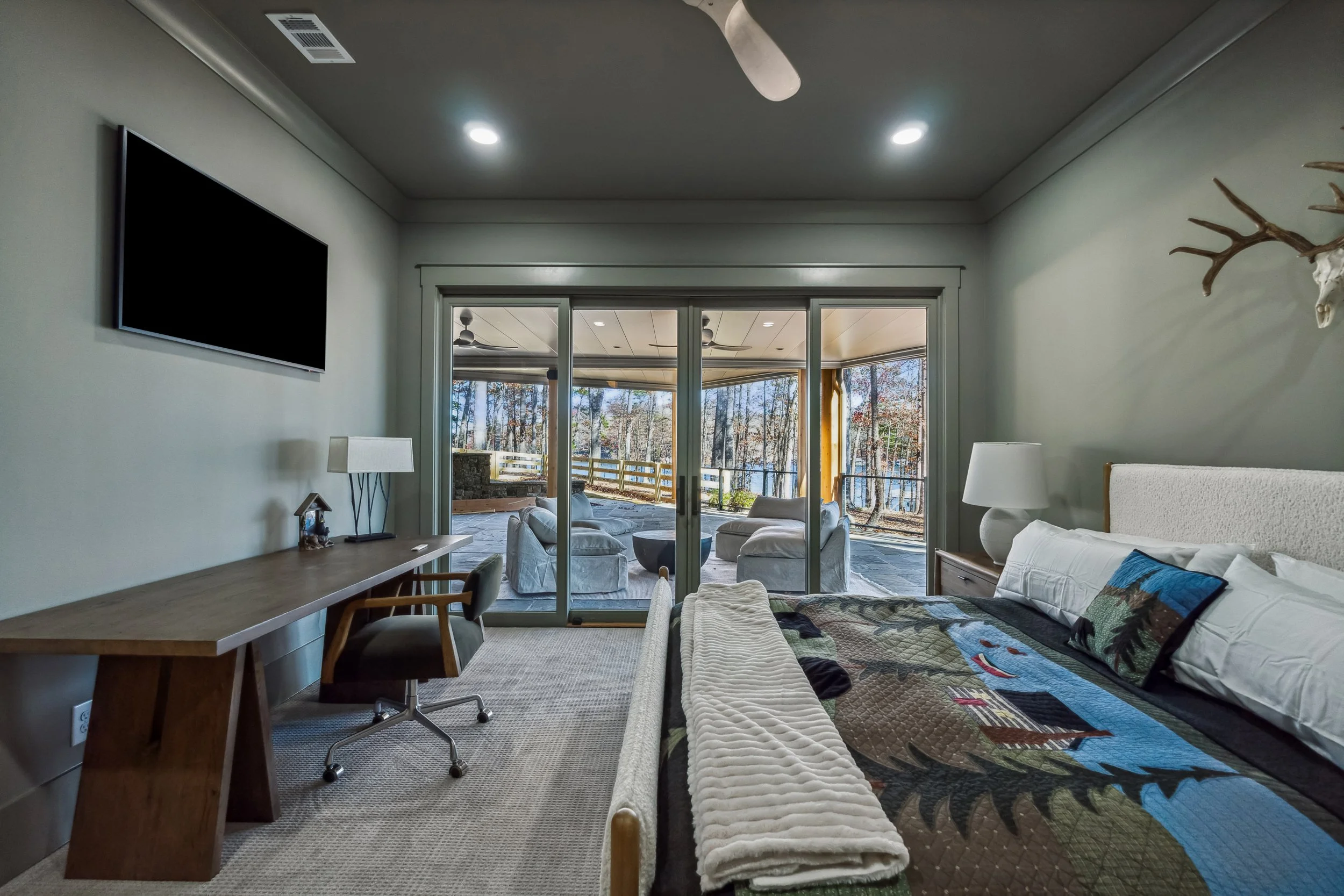 A bedroom with a large sliding glass door leading to a screened porch and a scenic view of trees and water. The room has a white bed with a quilt, a wooden nightstand with a lamp, and a mounted deer skull with antlers on the wall. There is a white be