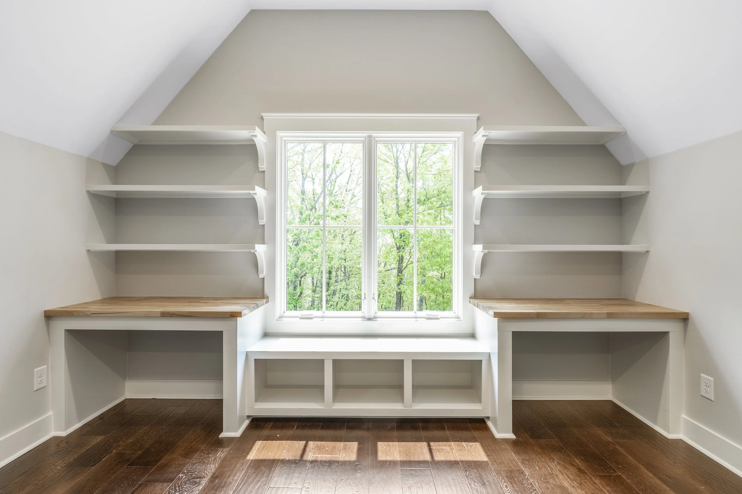 Empty room with large window and built-in white shelves on either side, hardwood floors, and a view of green trees outside.