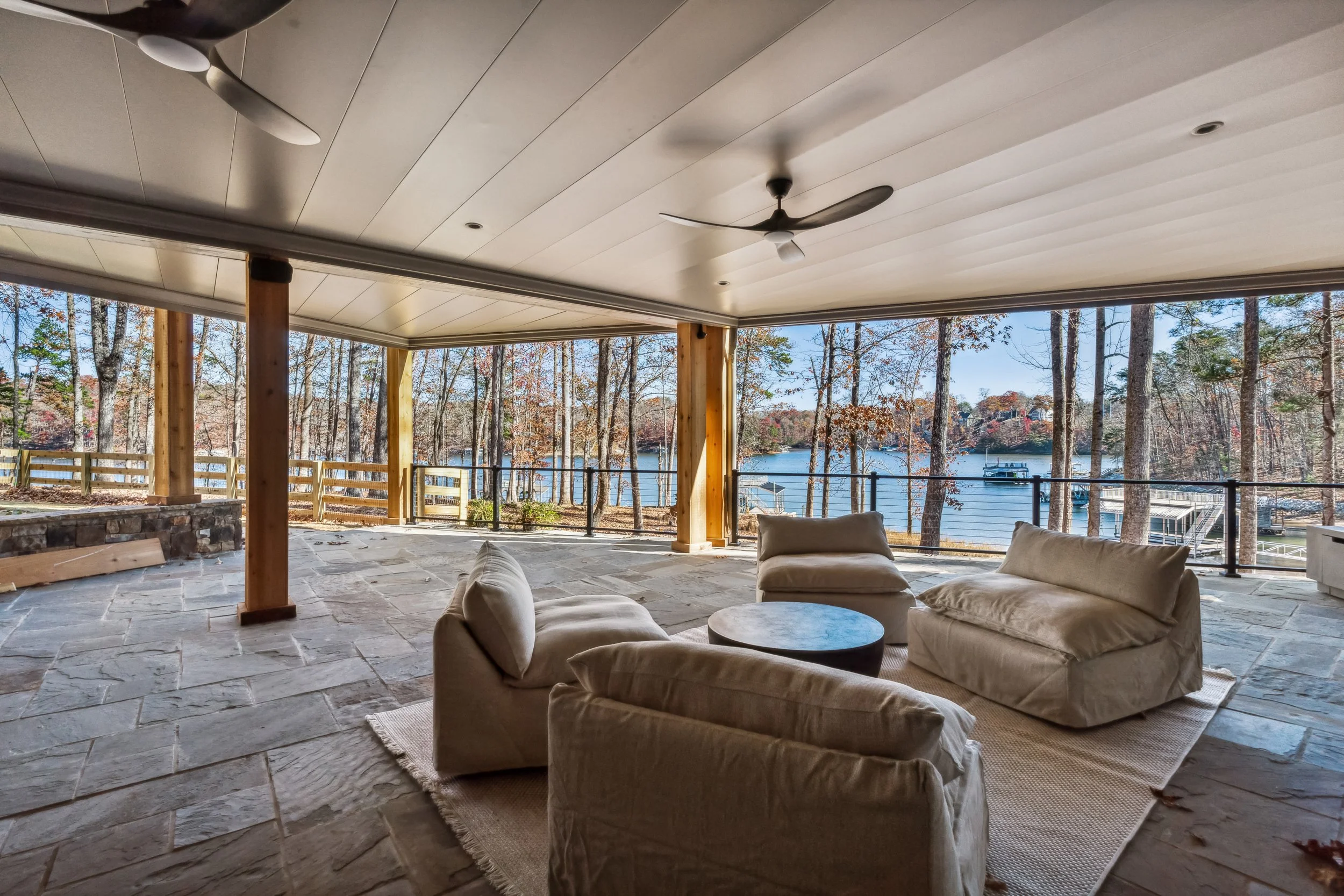 A covered outdoor patio with beige cushioned furniture, stone flooring, wooden posts, ceiling fans, and a view of a lake surrounded by trees with autumn foliage.