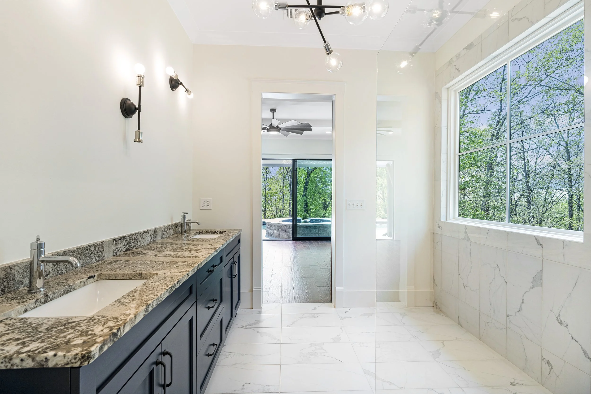 Modern bathroom with large window showing trees outside, granite countertop with two sinks, dark blue cabinetry, white marble floor and wall tiles, and black wall-mounted light fixtures.