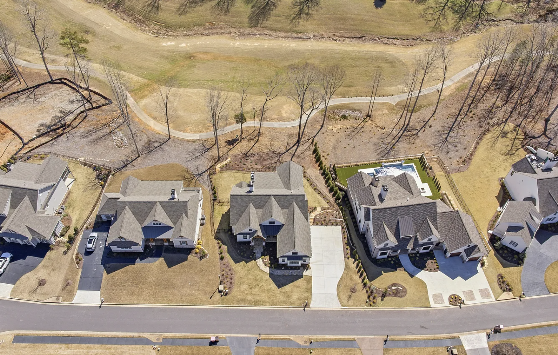 An aerial view of a residential neighborhood with several houses, yards, a small swimming pool, and a large open land area with trees and walking paths.