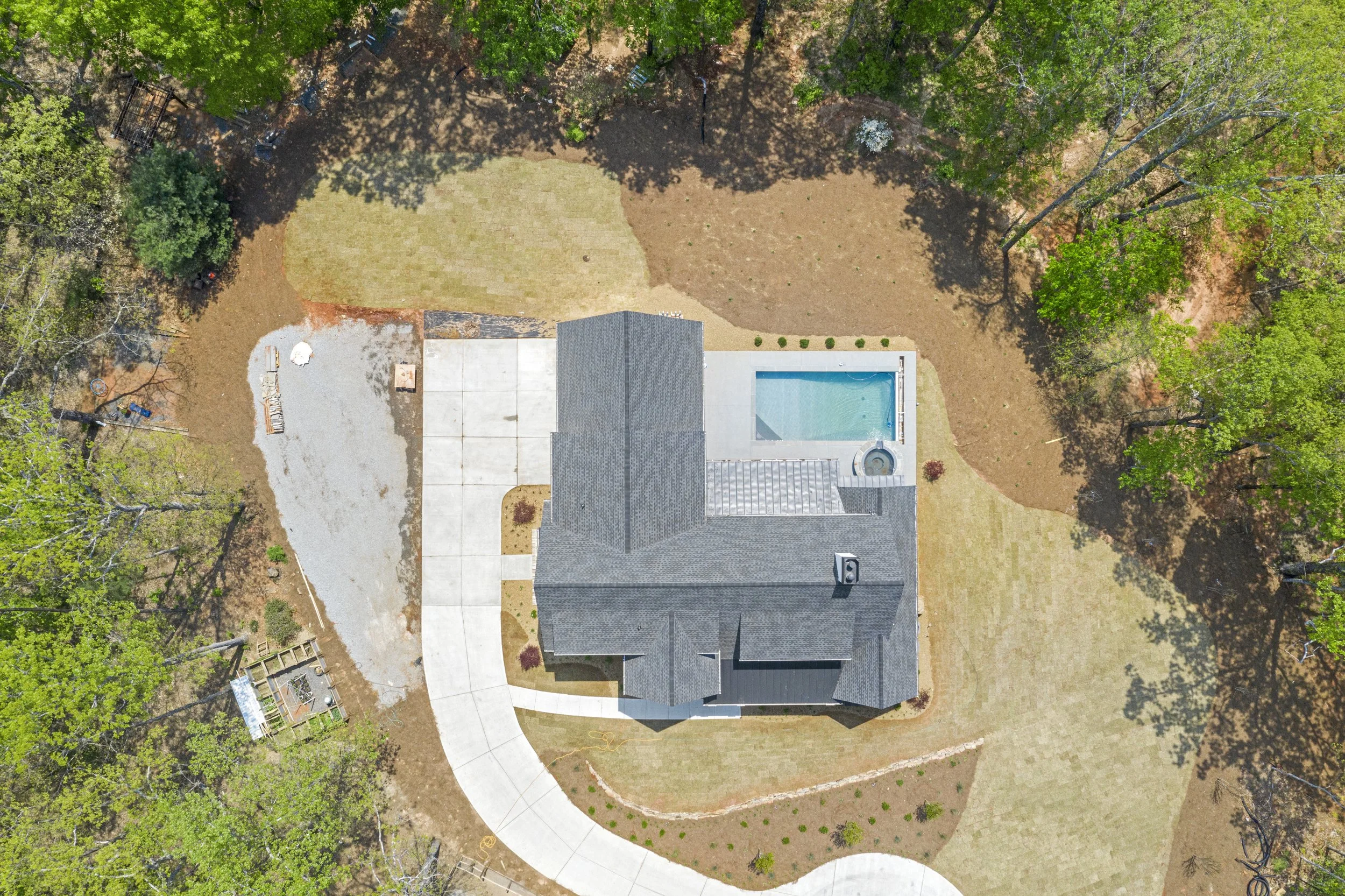 Aerial view of a house with a pool, driveway, and yard surrounded by trees.