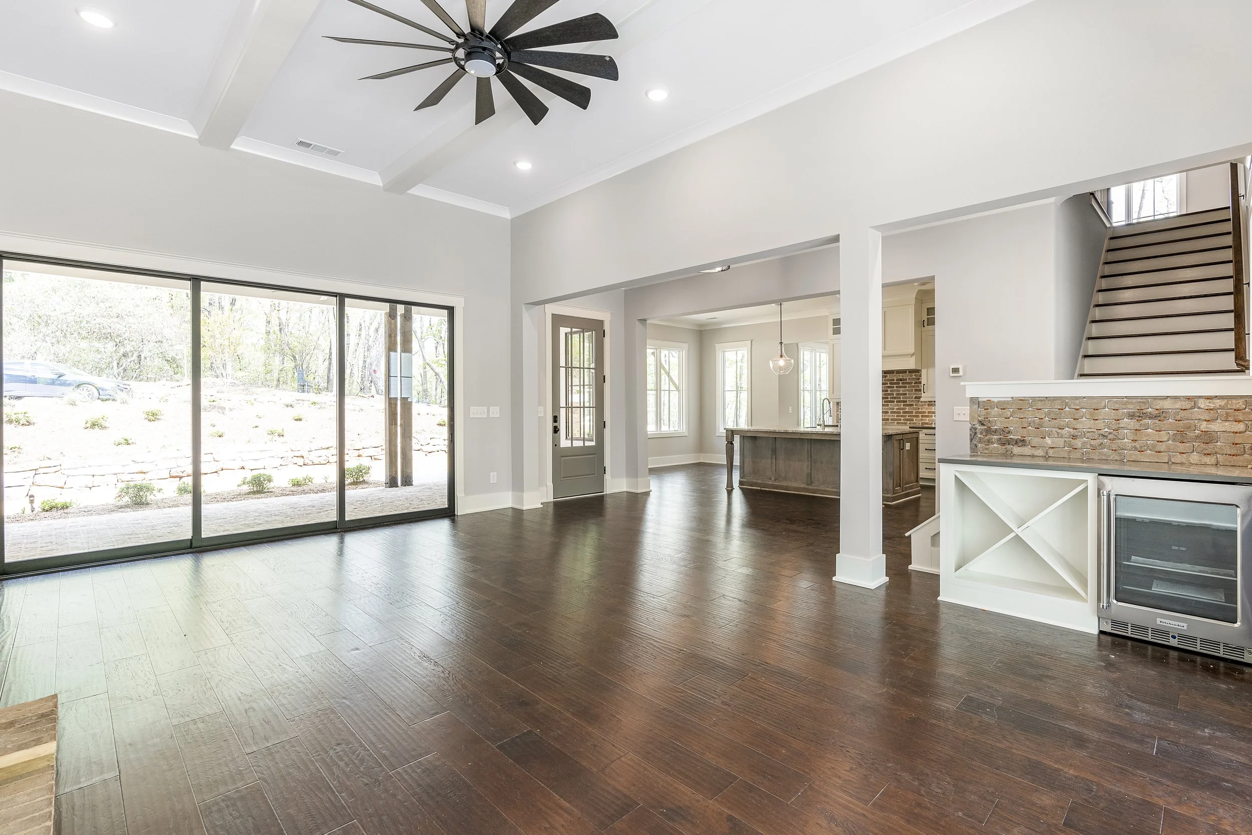 Interior of a modern, bright living space with large glass sliding doors, white walls, dark hardwood flooring, a ceiling fan, and some stairs leading to an upper level. The space connects to a kitchen area with a brick accent wall and a pendant light