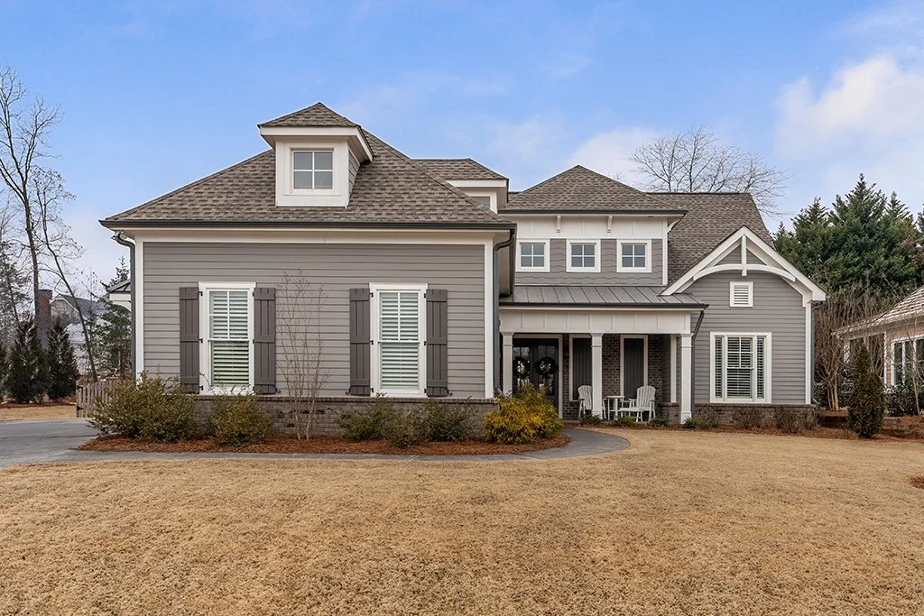 A large, two-story gray house with white trim, a front porch with chairs, and a well-maintained lawn.