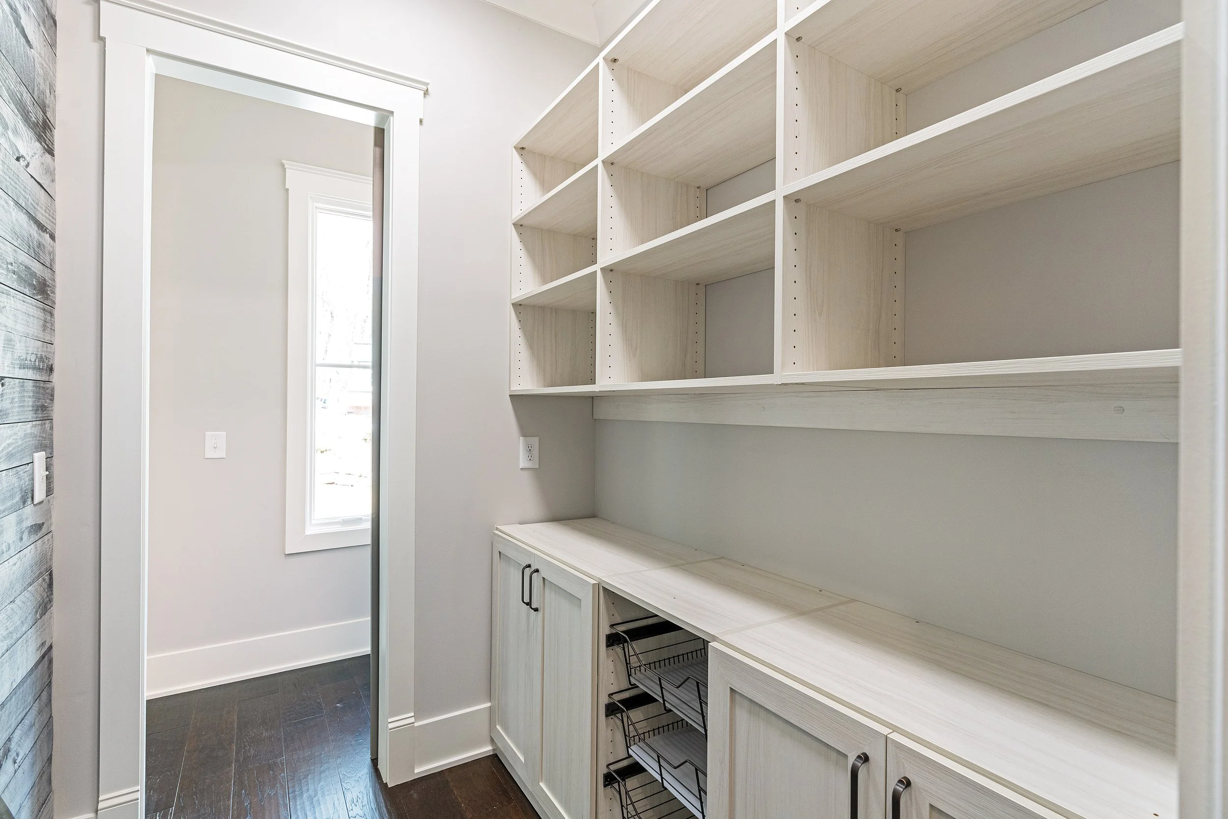 Empty white kitchen cabinets with open shelves and wire baskets, next to a white door and window, dark hardwood floors.