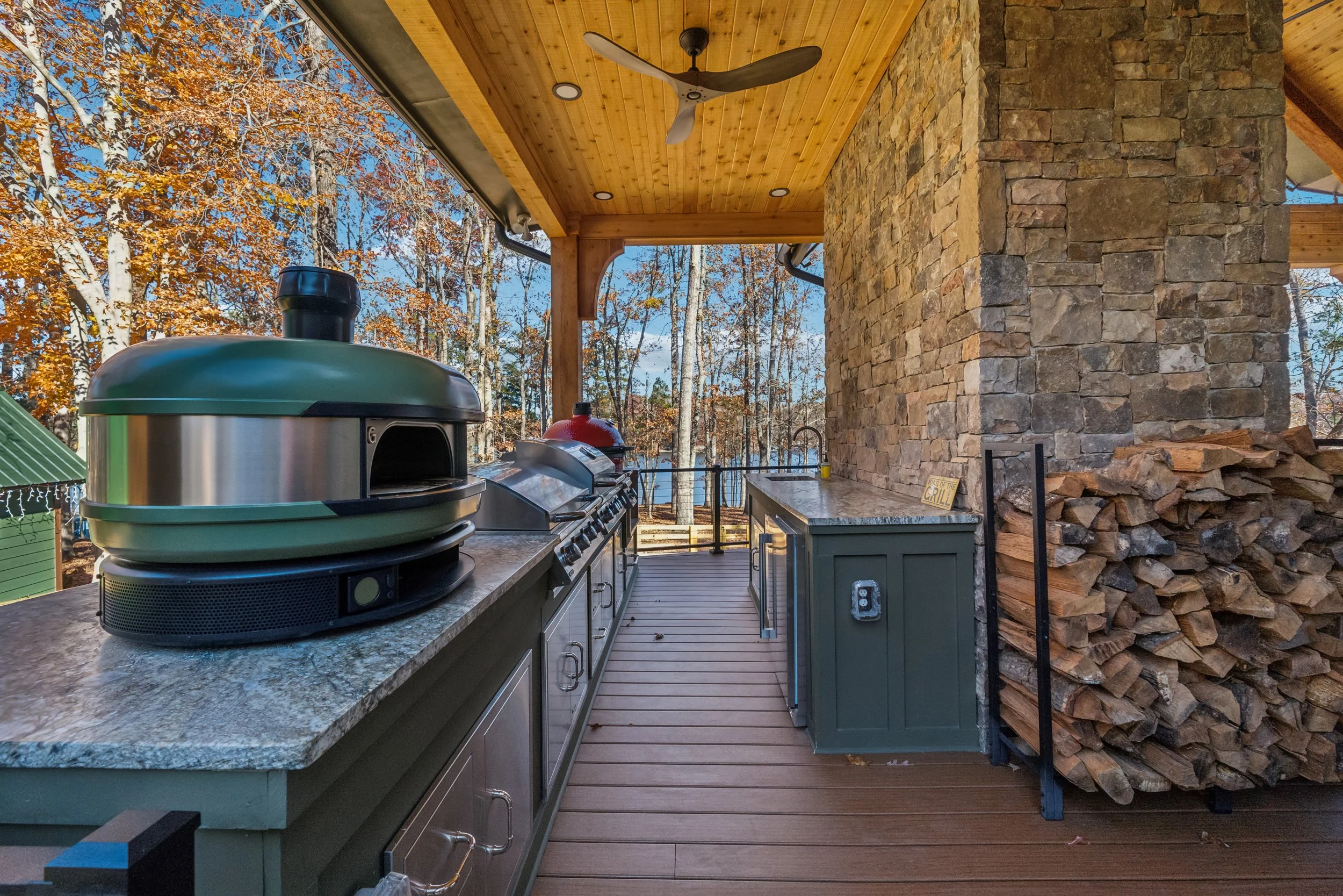 Outdoor kitchen with stone fireplace, wooden ceiling, ceiling fan, grill, and a view of autumn trees and a lake in the background.