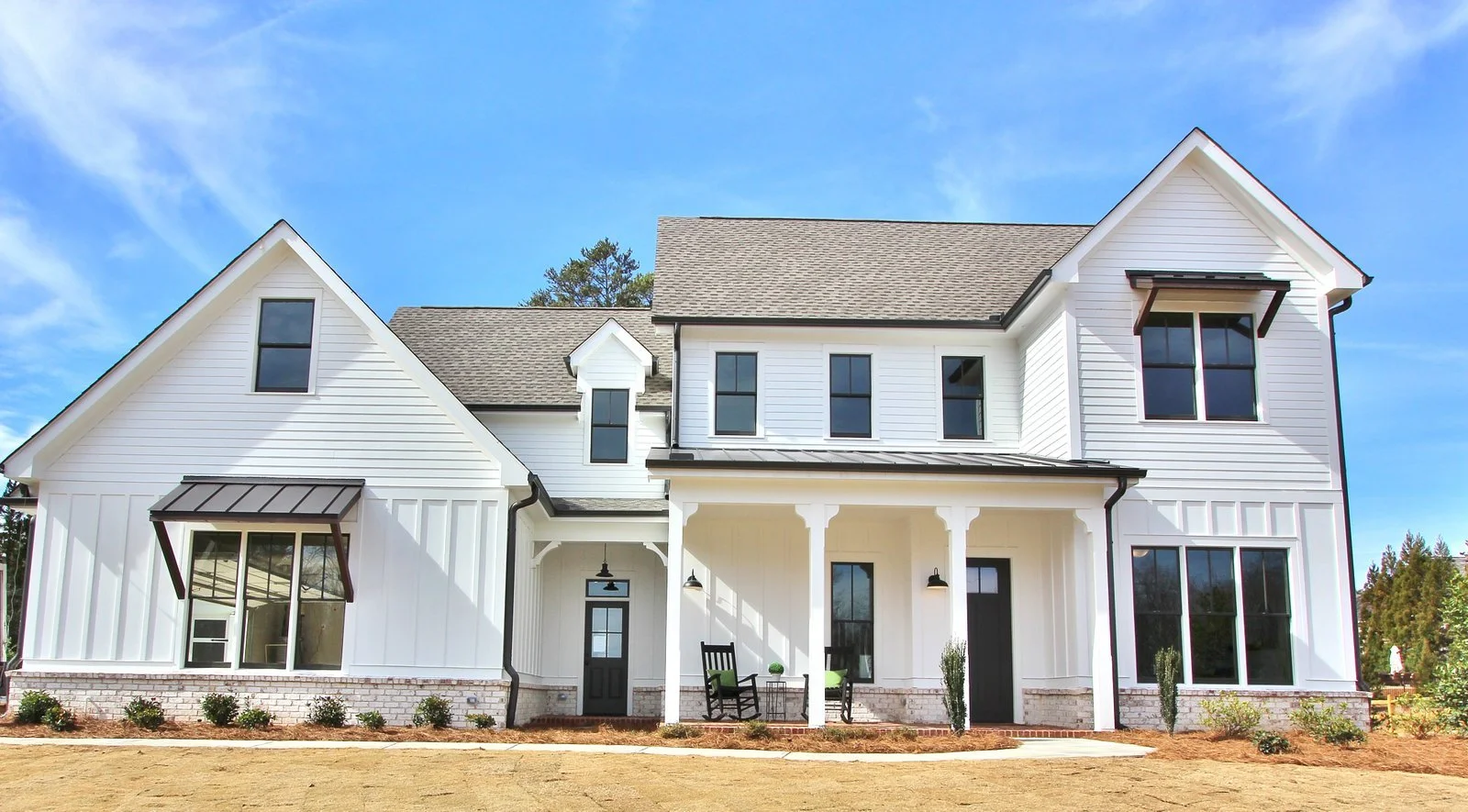 Modern two-story white house with black accents, porch seating, black window frames, and a gable roof, under a blue sky.