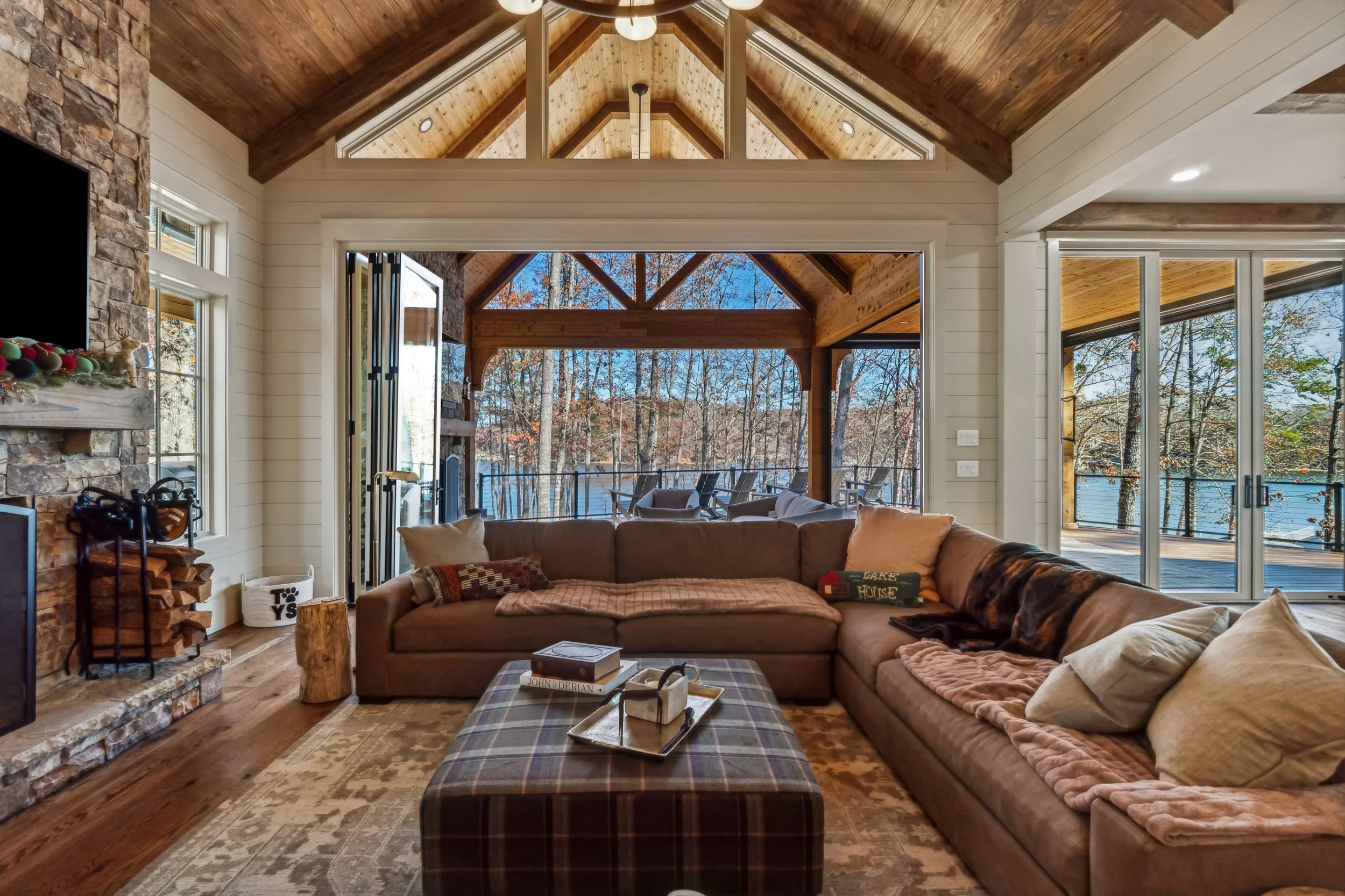 Interior view of a cozy living room with a vaulted wooden ceiling, large windows showing a wooded outdoor scene, a stone fireplace, and comfortable seating including a large sectional sofa and an ottoman.