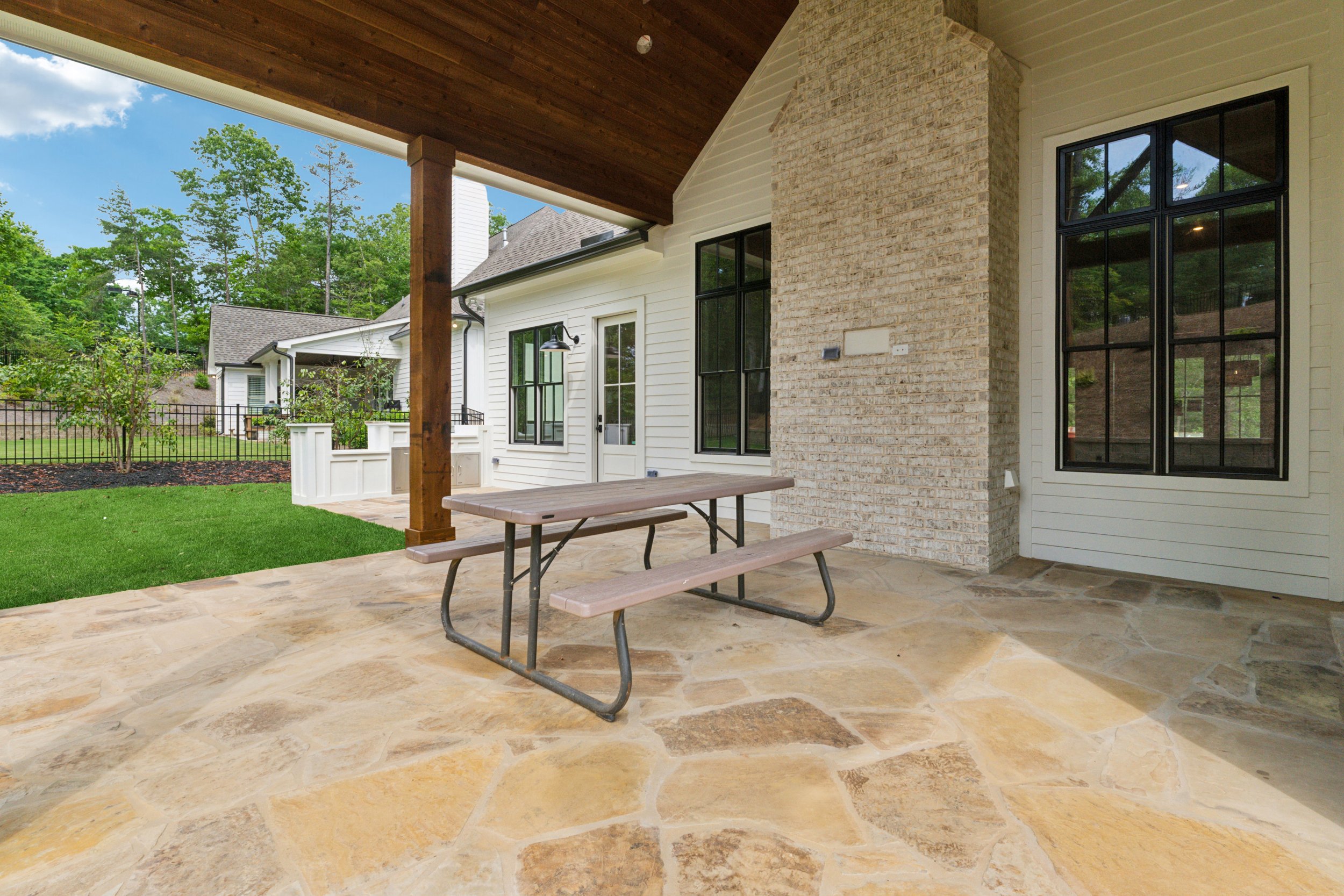 Covered patio area with stone floor, wooden ceiling, picnic table, and view of backyard with green lawn and trees.