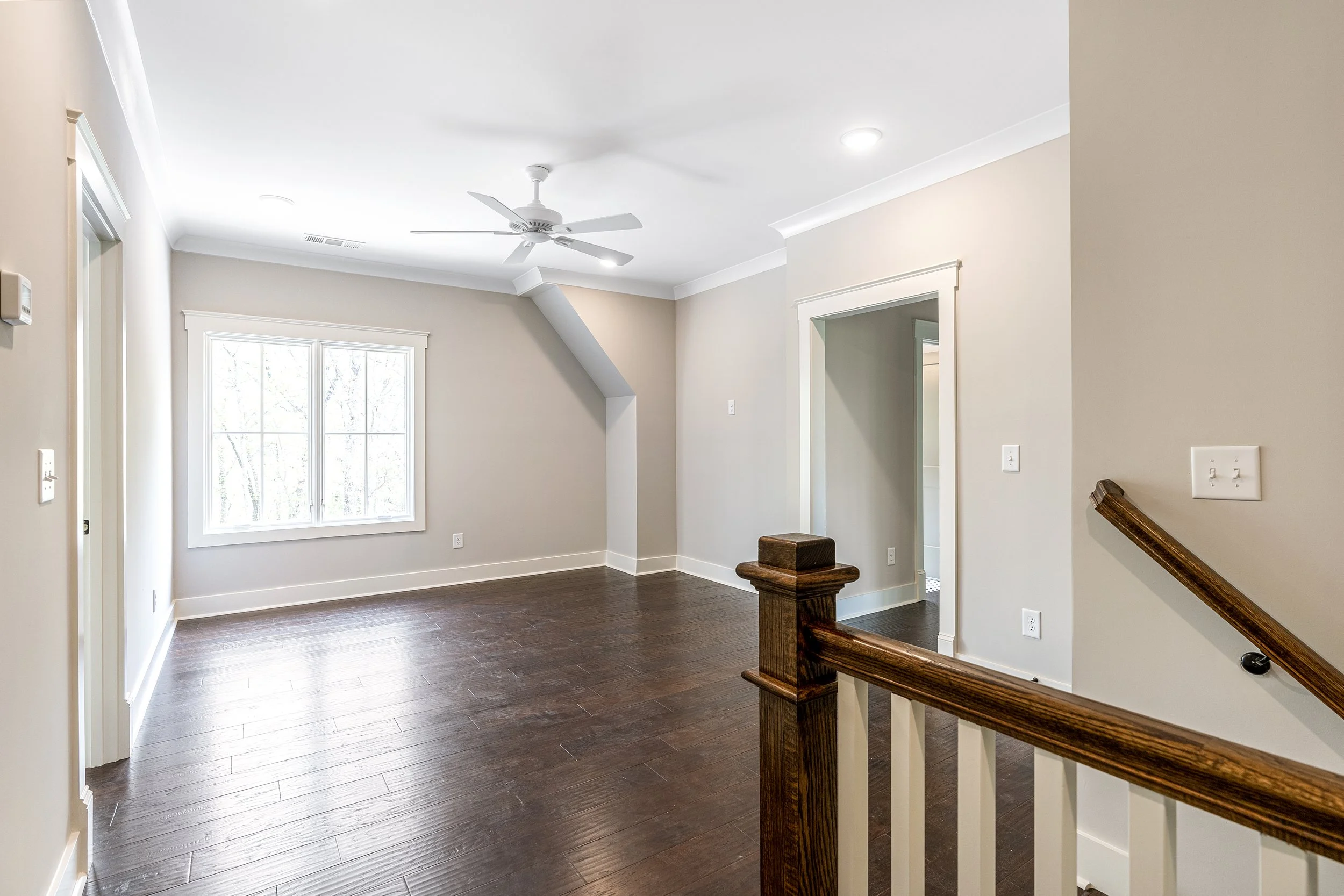 Interior view of a modern home with dark wooden flooring, white walls, large window, ceiling fan, classic trim, and stairs with a wooden handrail.
