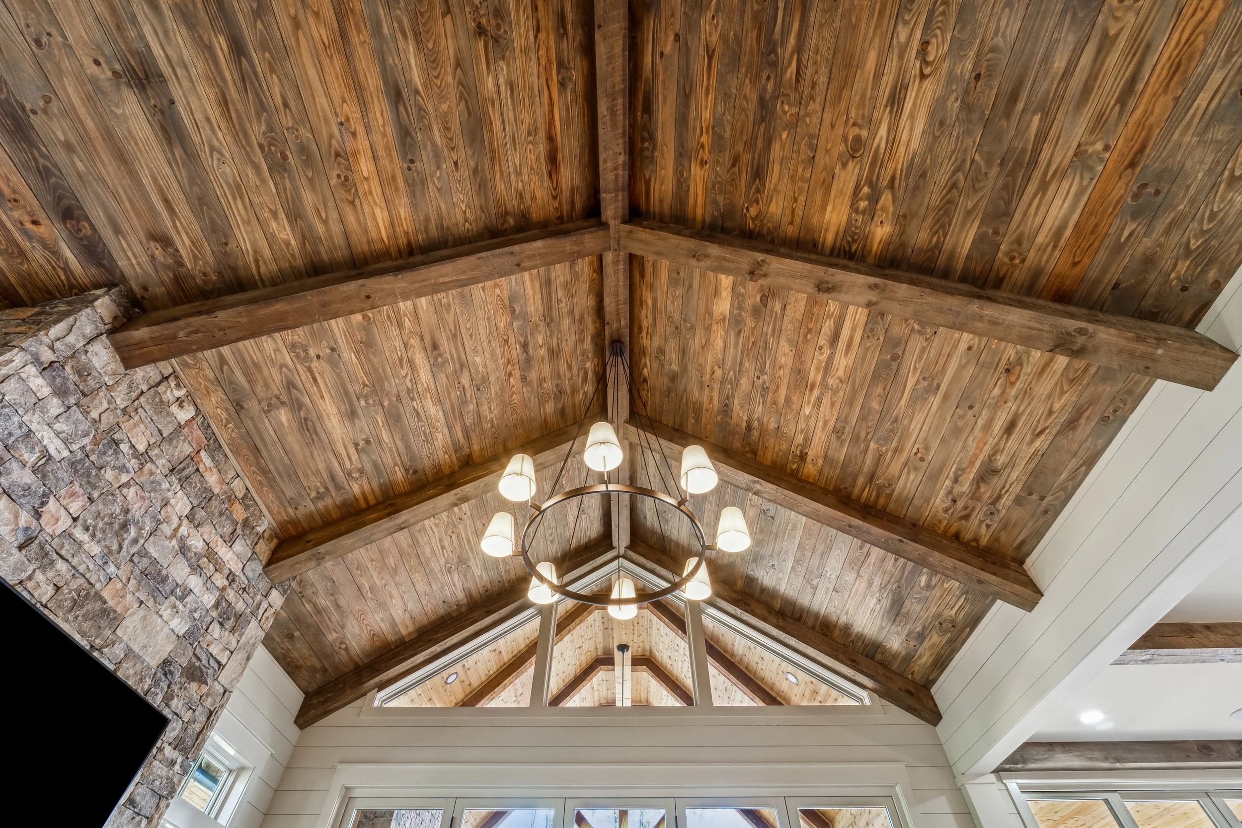 Interior view of a wooden ceiling with exposed beams and a central chandelier with multiple lights in a room with large windows.