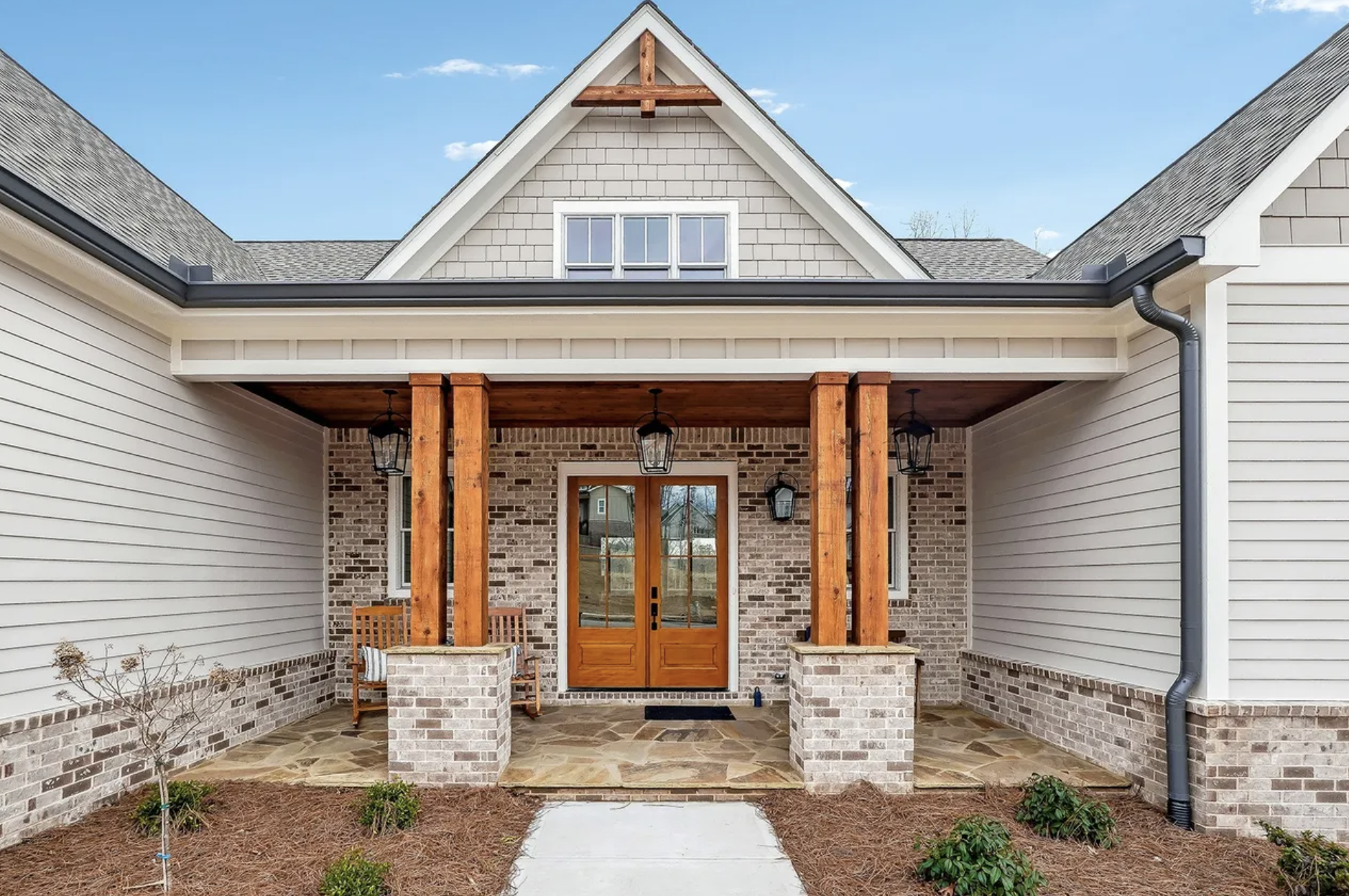 Front porch of a house with brick, siding, and wood accents including a wooden door and porch posts, two black outdoor lanterns, small plants, and a stone pathway.