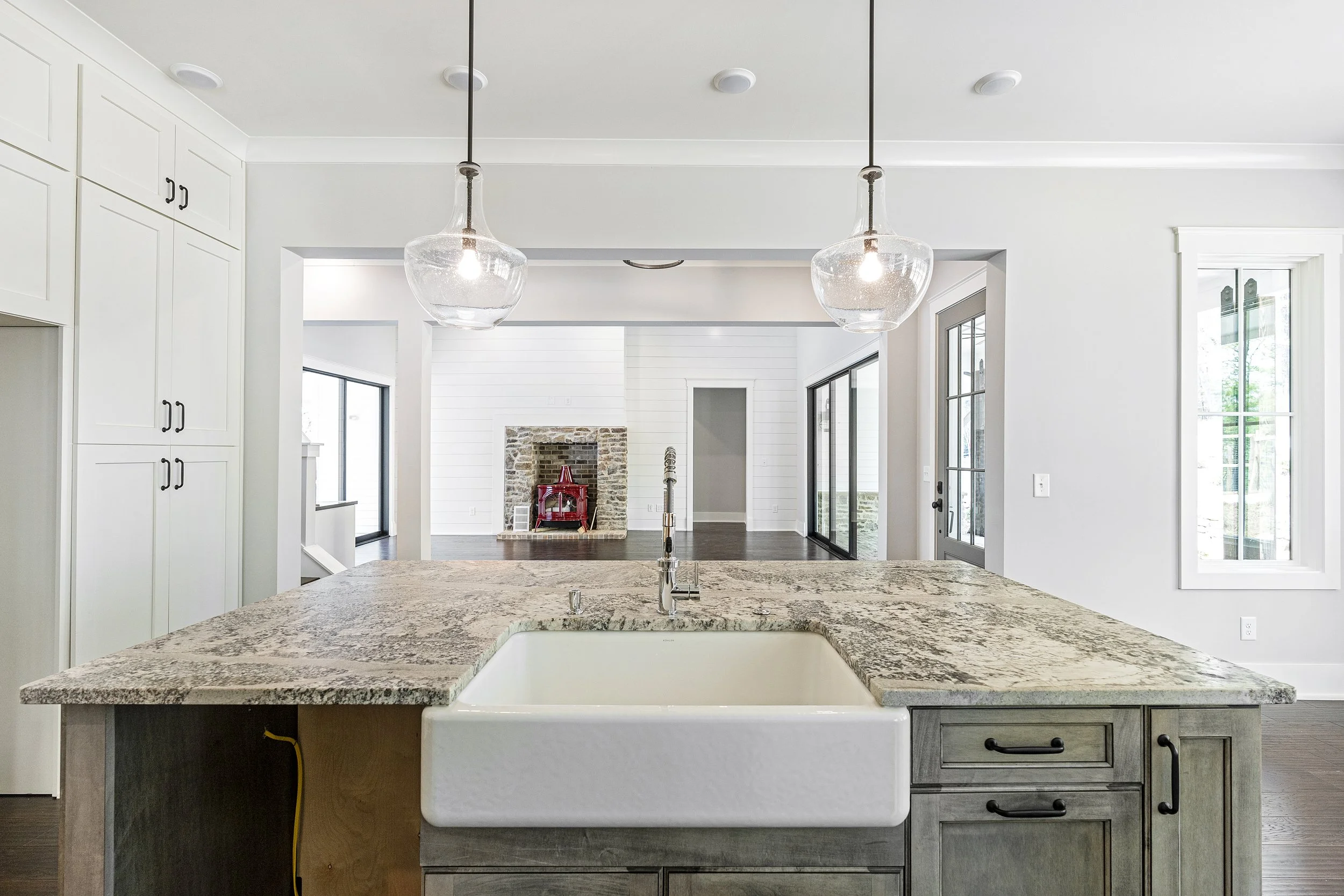 A modern kitchen with a granite island counter, a white farmhouse sink, and black cabinetry. Two glass pendant lights hang from the ceiling above the island. In the background, a living area with a brick fireplace and a red decorative piece is visibl