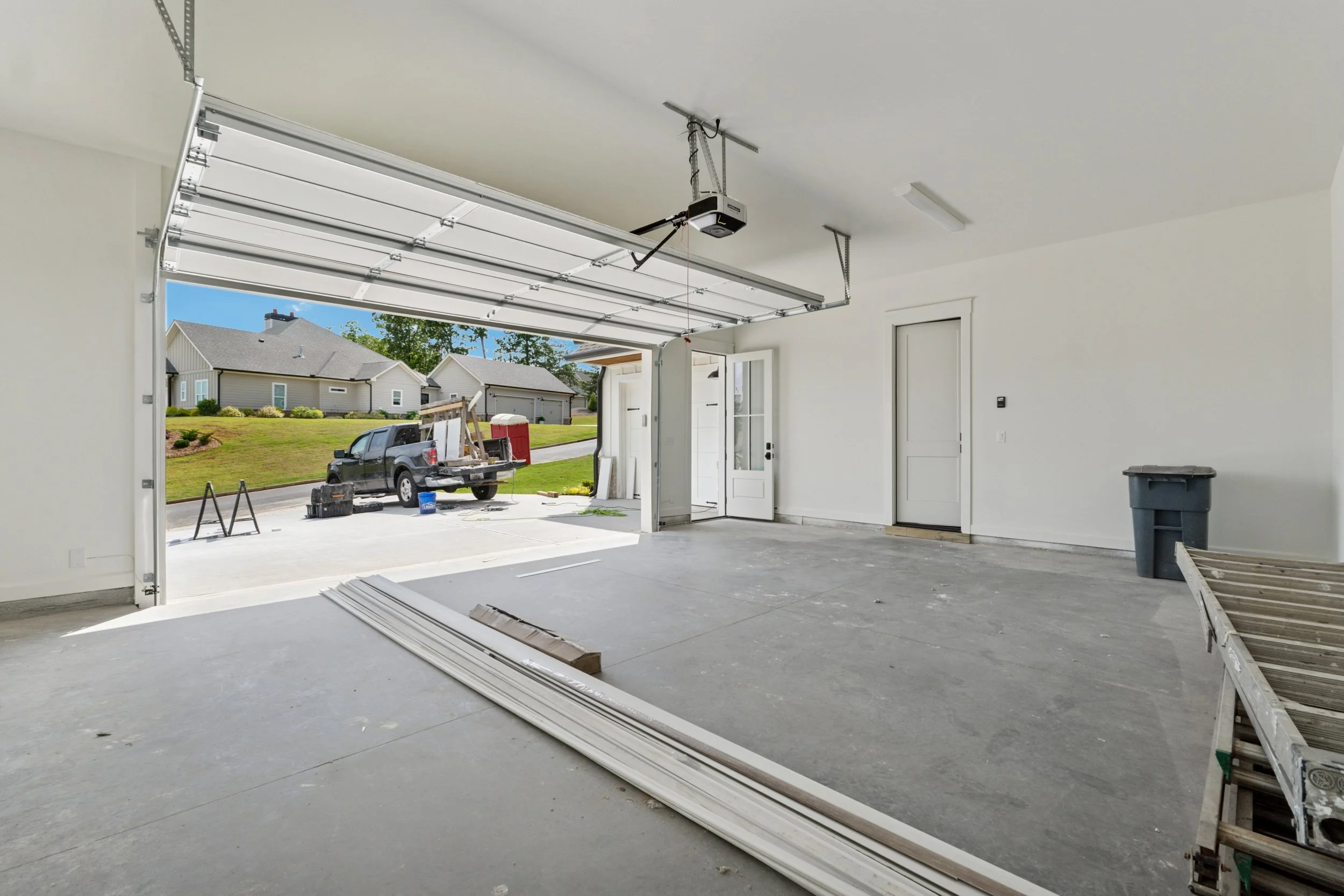 Interior of a house garage with an open door, construction tools, and materials, with residential neighborhood visible outside.