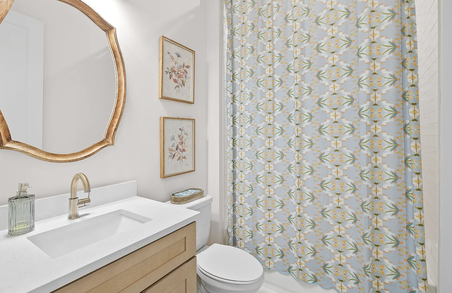 A bathroom with a white sink, light brown vanity, and a large mirror with a wooden frame. Two framed floral pictures hang on the white wall. A soap dispenser is on the sink, and a small basket with tissue is on the counter. A toilet is next to the si