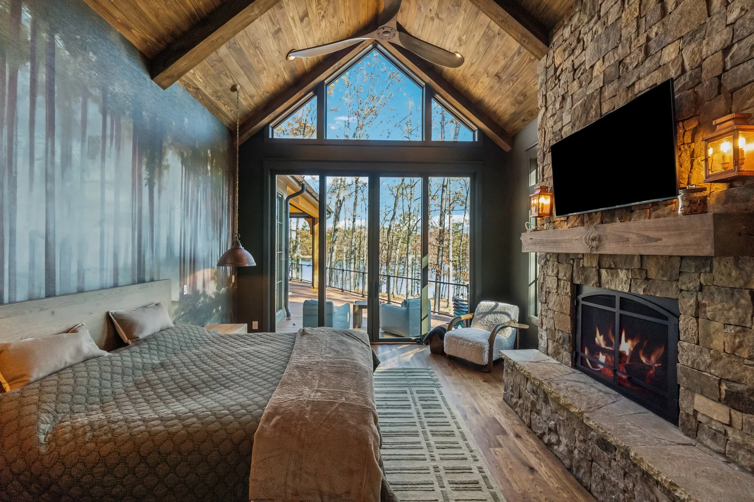Living room with a vaulted wooden ceiling, large windows showing trees outside, a stone fireplace with a mounted TV, and a bed with pillows and quilted blankets.