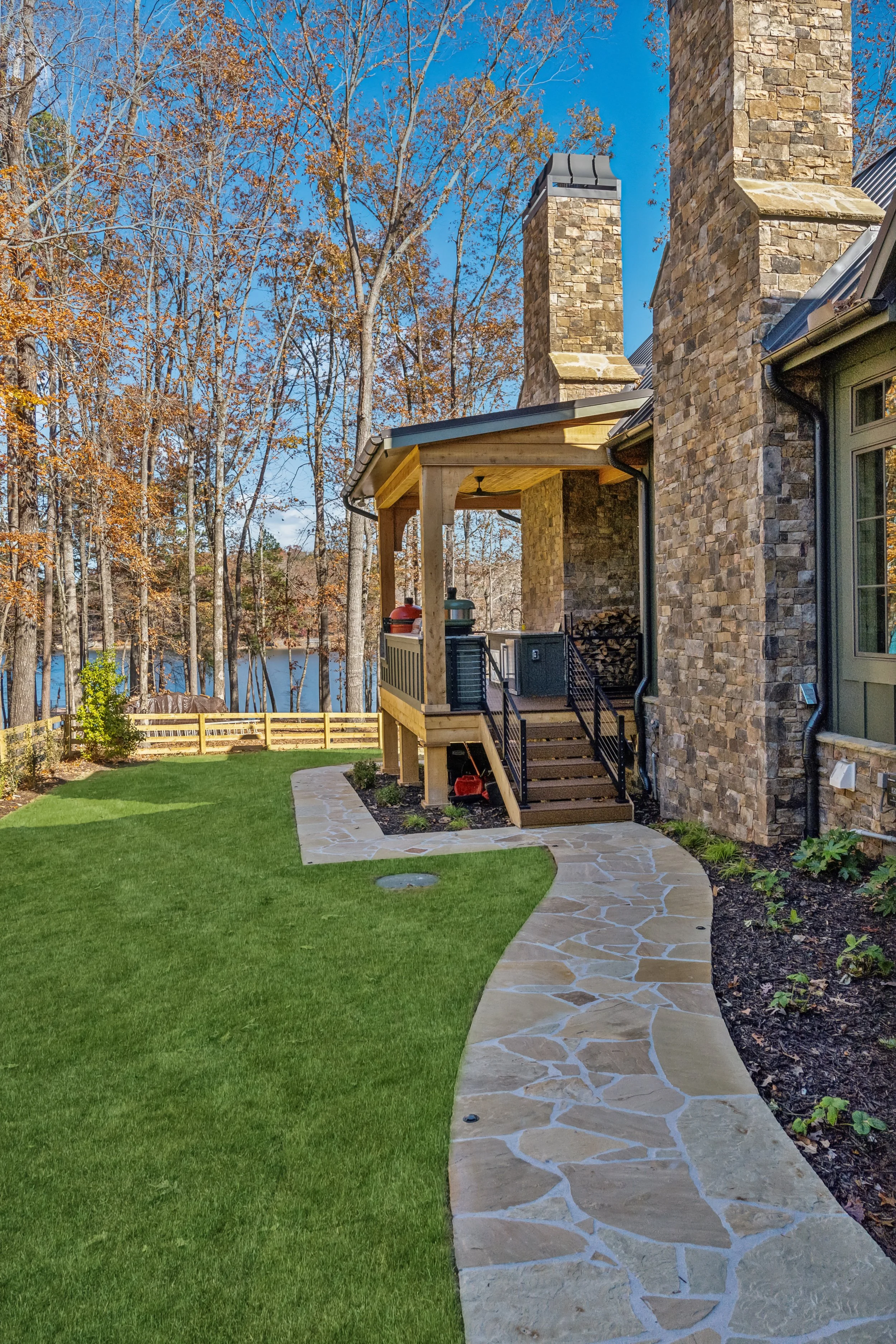 A backyard scene with a stone house featuring multiple chimneys, a wooden deck with grills, a manicured lawn, a stone pathway, and trees with fall foliage.