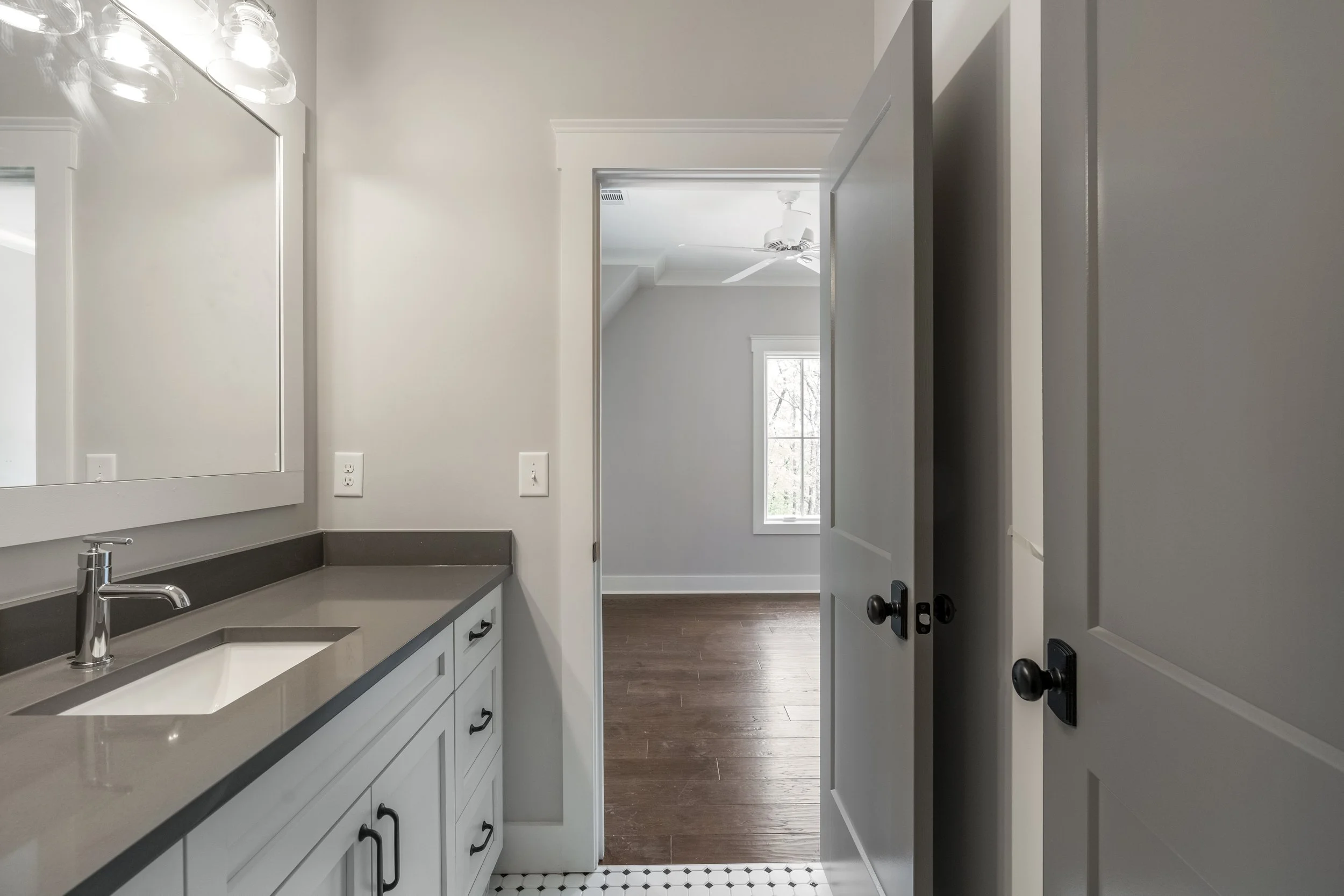 A bathroom vanity with white cabinets and a dark gray countertop, a faucet, and a large mirror. A door is open, revealing a room with a ceiling fan and a window with natural light.