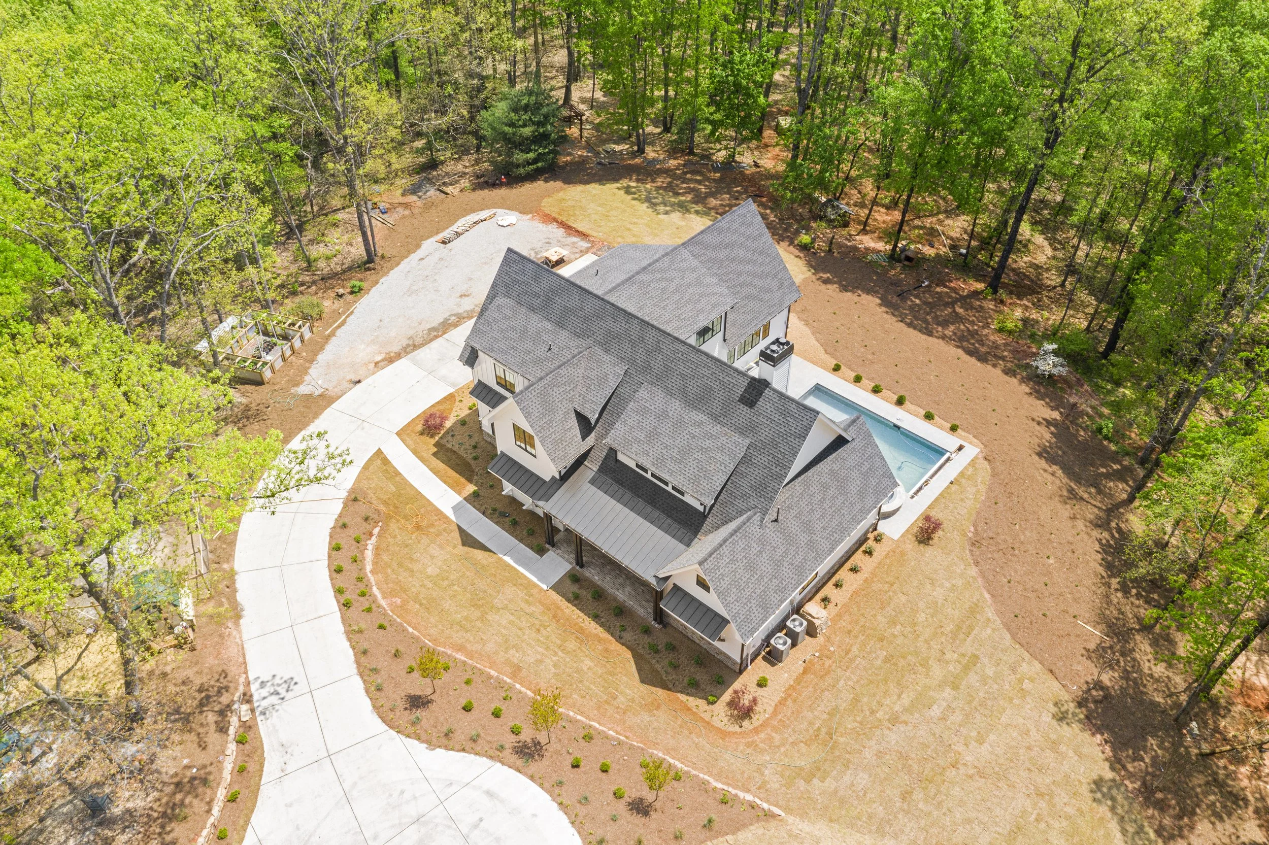 Aerial view of a house surrounded by trees, with a driveway, a backyard pool, and a landscaped yard under sunny weather.