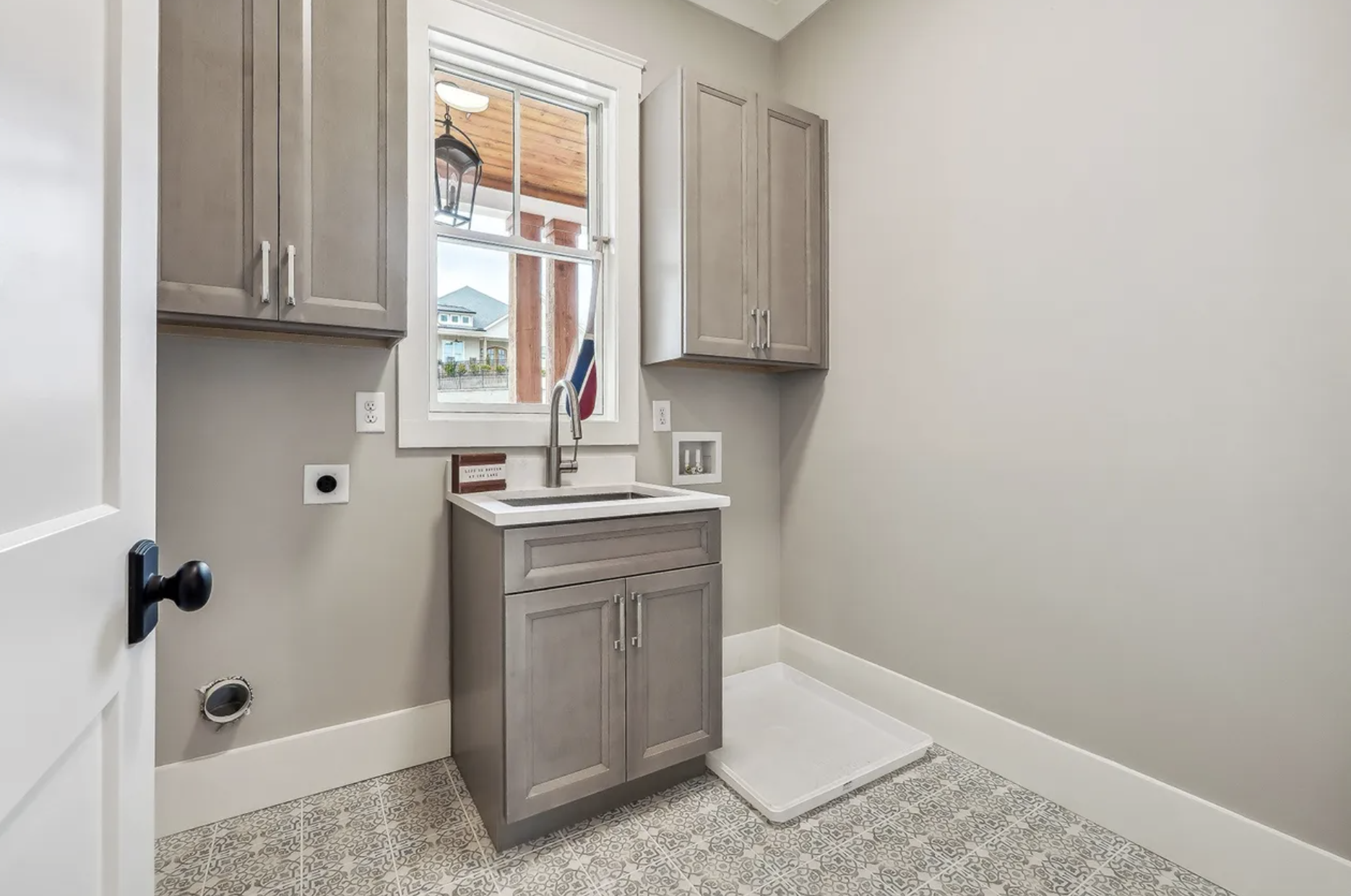 A laundry room with a window, kitchen-style cabinets, a countertop with a sink, and decorative patterned floor tiles.