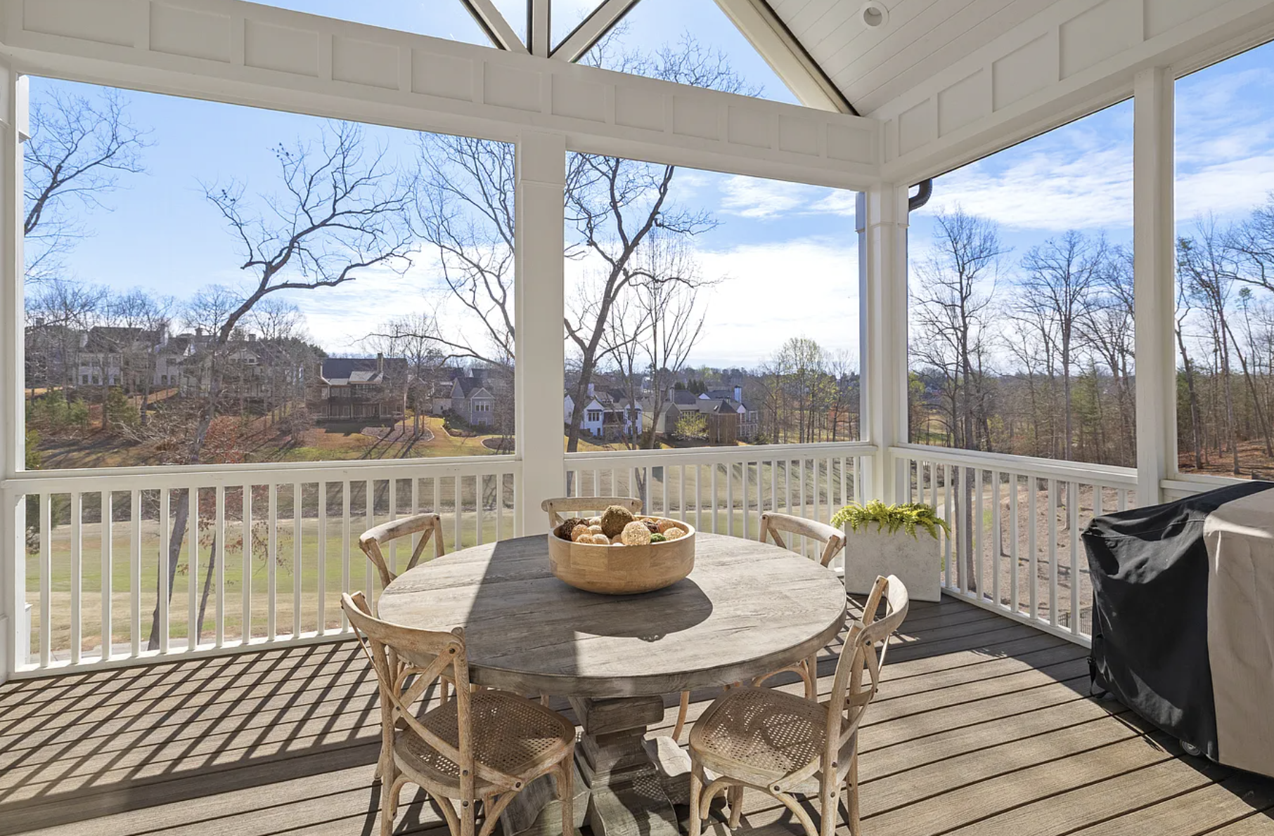 View from a screened porch with a round wooden table surrounded by six chairs, a bowl of decorative objects in the center, a potted plant, and a covered grill. The background shows leafless trees and houses under a partly cloudy sky.