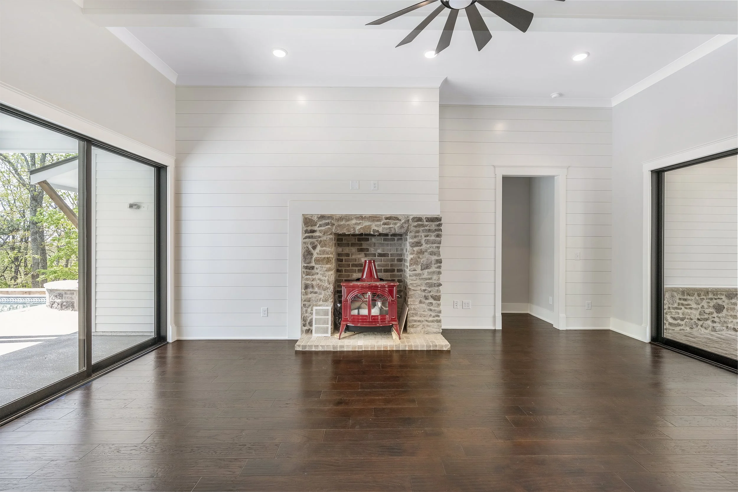Living room with white walls, a stone fireplace with a red decor item on the hearth, dark wood flooring, sliding glass doors leading outside, and a ceiling fan.