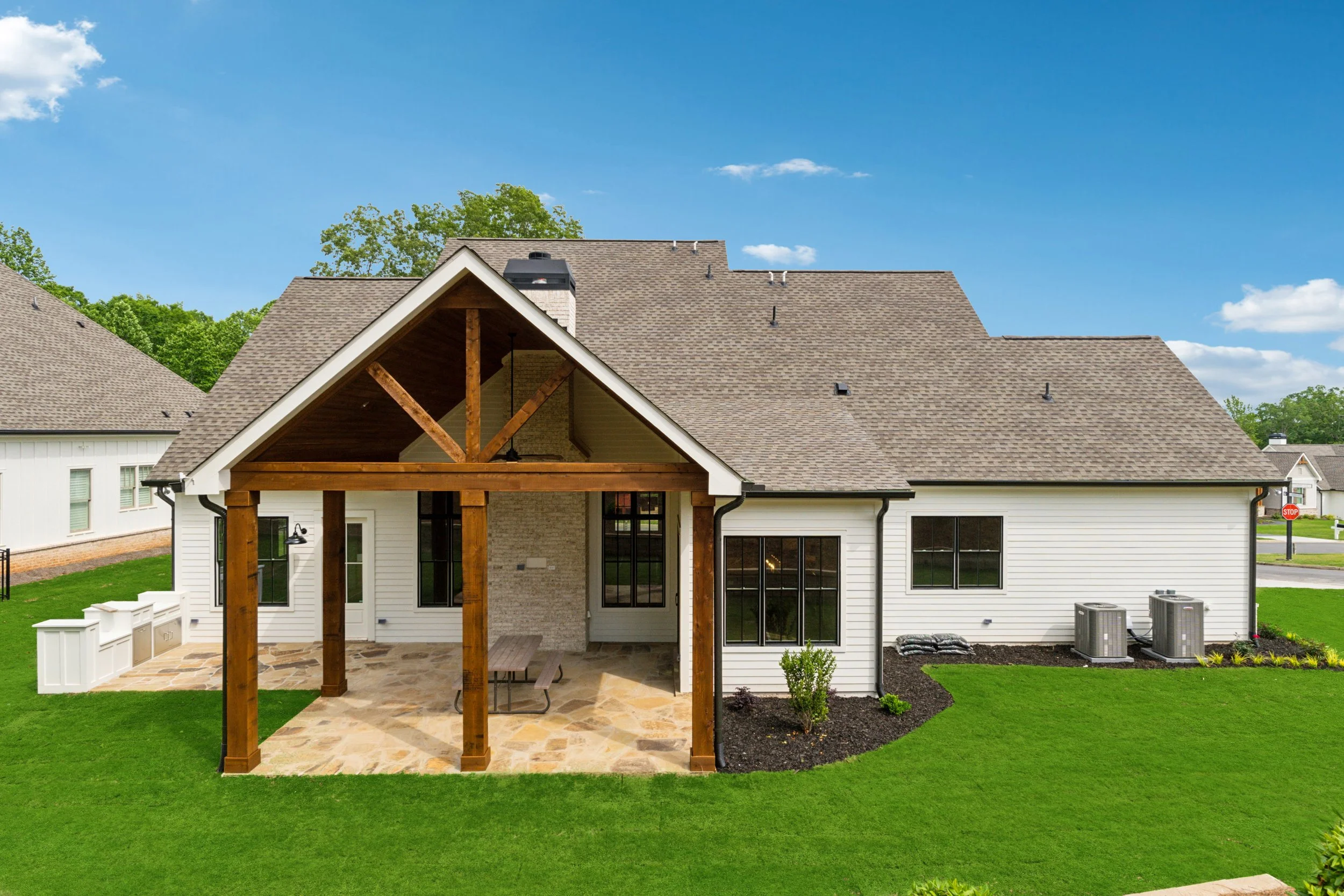 Rear view of a white house with a wooden patio covering, stone flooring, black-framed windows, and a well-maintained green lawn, under a blue sky with some clouds.