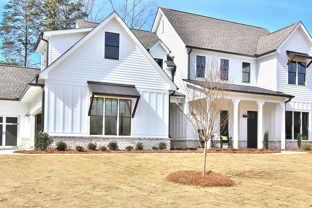 A large modern white house with a gabled roof, multiple windows, and a porch; a young leafless tree in front and some small bushes along the house foundation; dry grass in the front yard; a clear blue sky and trees in the background.