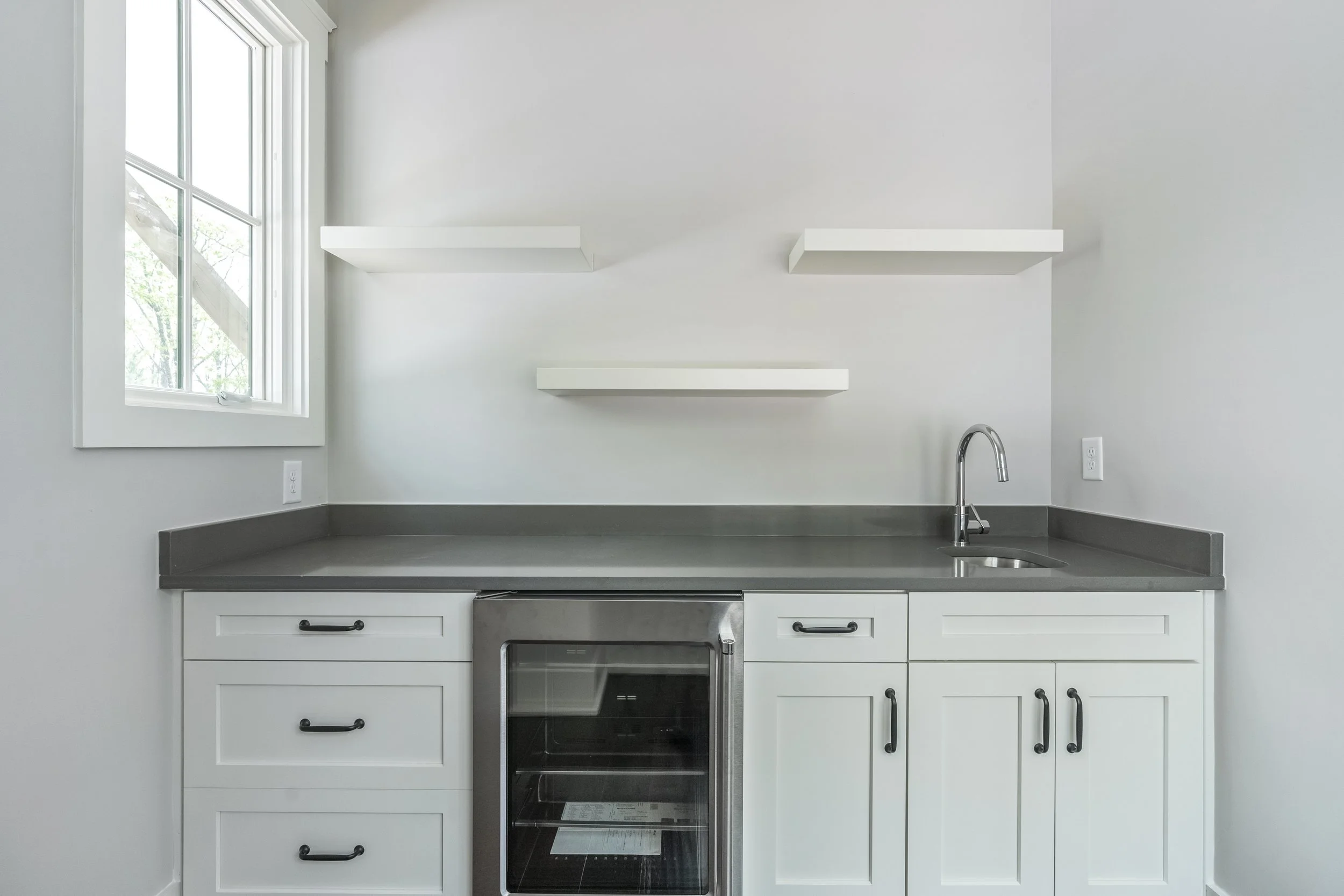 A minimalist kitchen with white cabinets, a gray countertop, a built-in oven, a stainless steel sink, and three white floating shelves on a white wall near a window.
