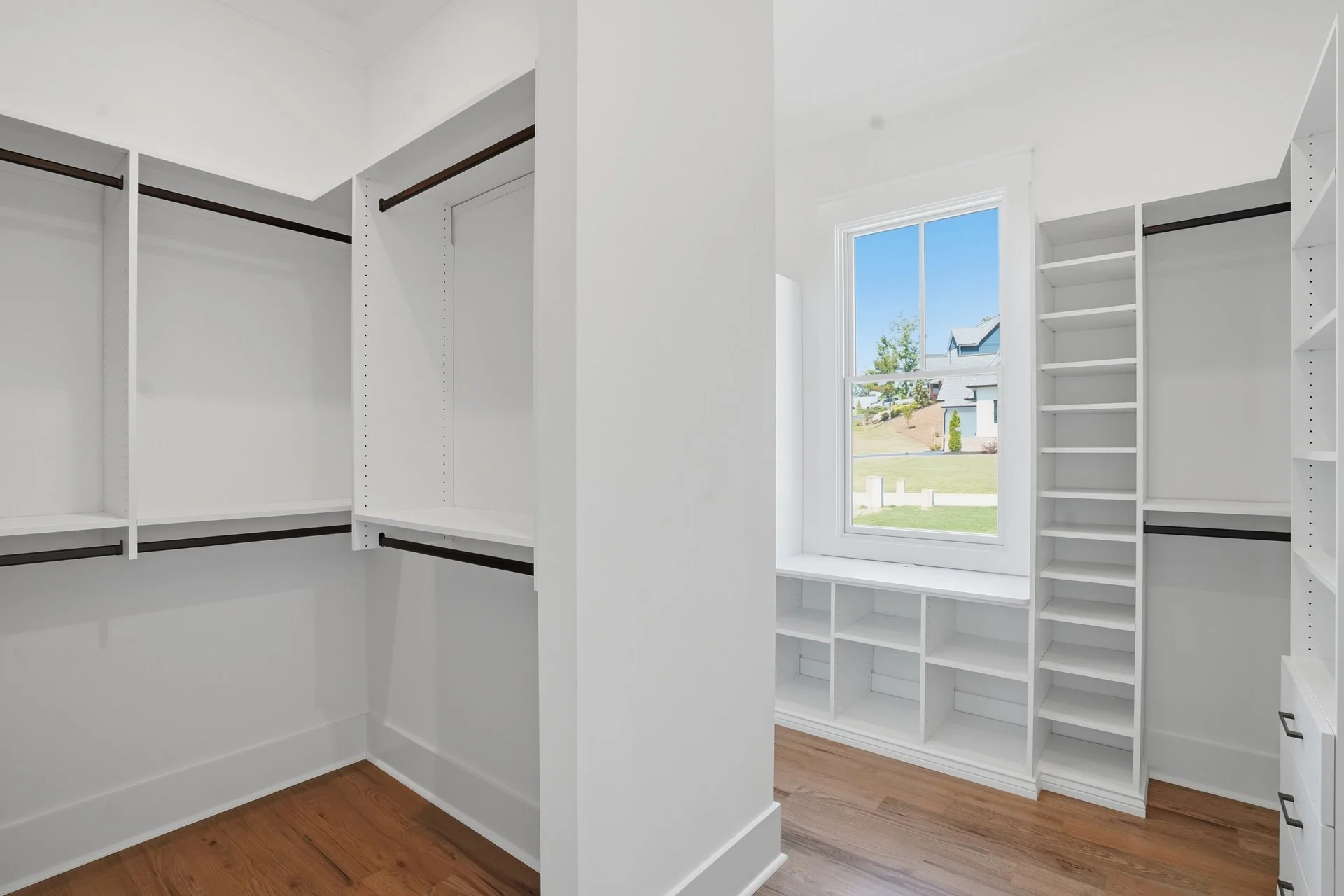 Empty white walk-in closet with built-in shelves and clothing rods, a window showing a suburban neighborhood with houses and blue sky.