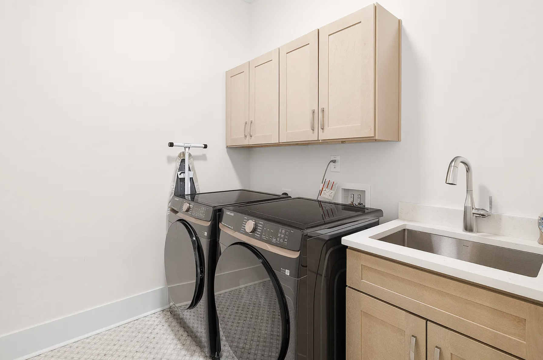 Laundry room with beige cabinets, a stainless steel sink and faucet, a Samsung washer and dryer, and an iron board against the wall.