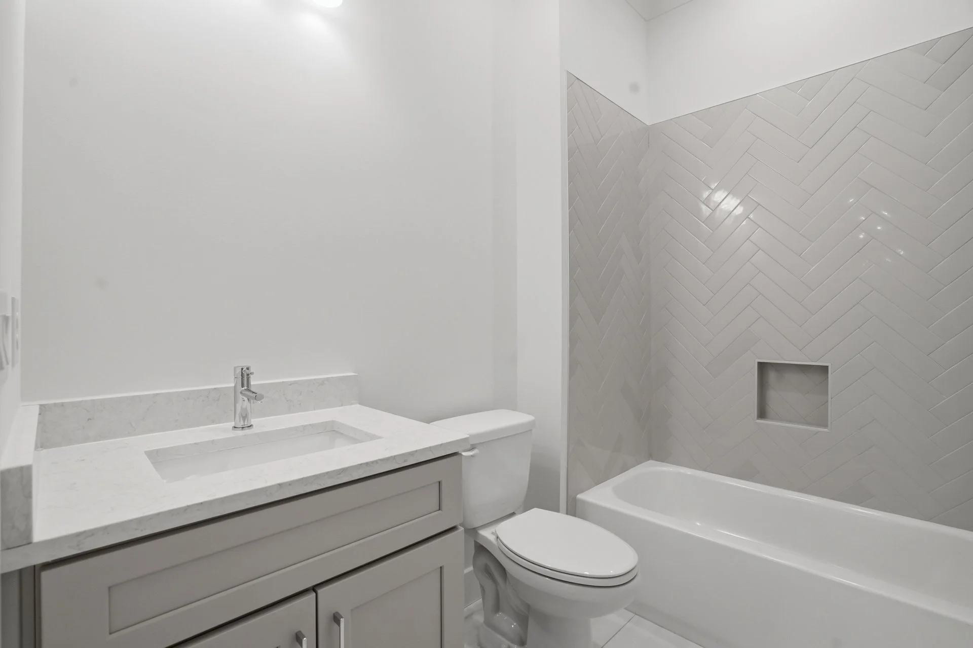 A modern bathroom featuring a white vanity with a marble countertop and a built-in sink, a white toilet, and a bathtub with a tile surround. The tiles on the shower wall are arranged in a herringbone pattern and have a glossy finish. The space is min