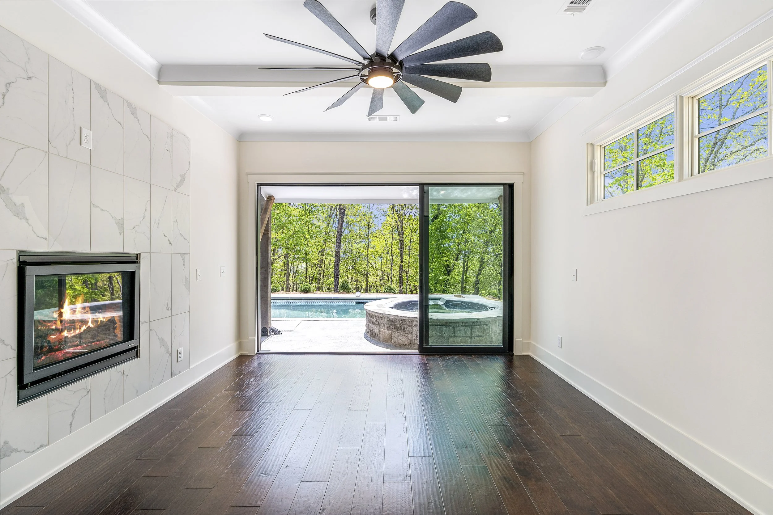 Interior of a modern living room with a ceiling fan, a fireplace, and large sliding glass doors opening to an outdoor pool and lush green trees.