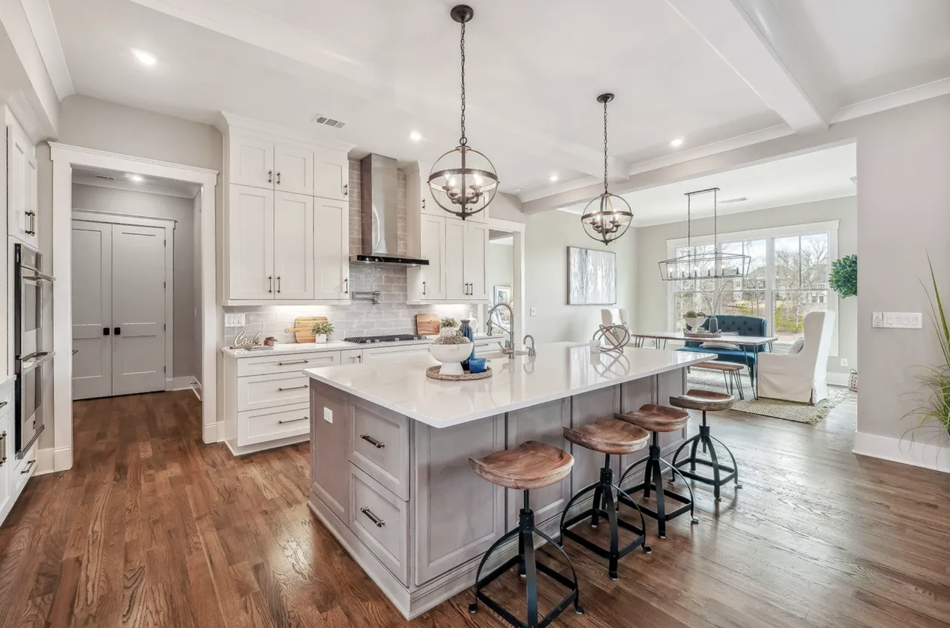 Open-concept kitchen and dining area with white cabinetry, a large island, pendant and chandelier lighting, hardwood floors, and large windows with a view of the backyard.